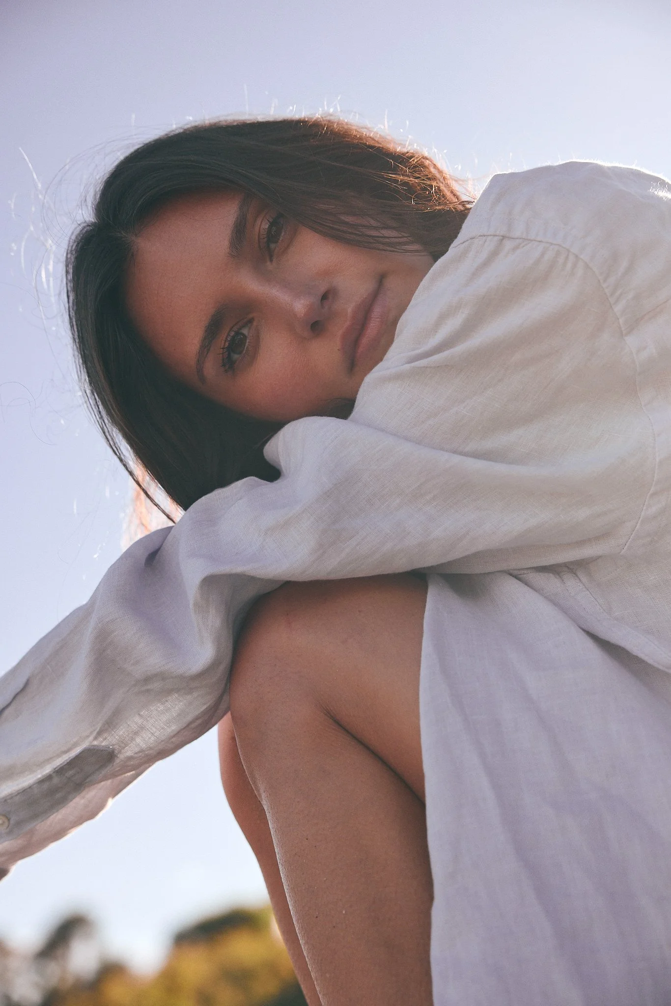 A woman with dark hair and a light complexion, smiling softly, leaning on her arm, wearing a loose white shirt, outdoors during daylight with blurred natural background.