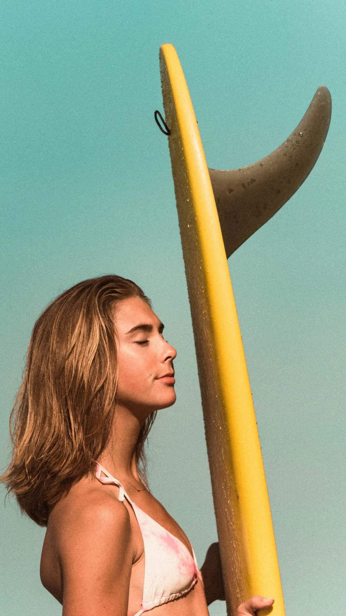 A woman with long, wavy hair and closed eyes standing with her side profile next to a yellow surfboard, against a clear blue sky.