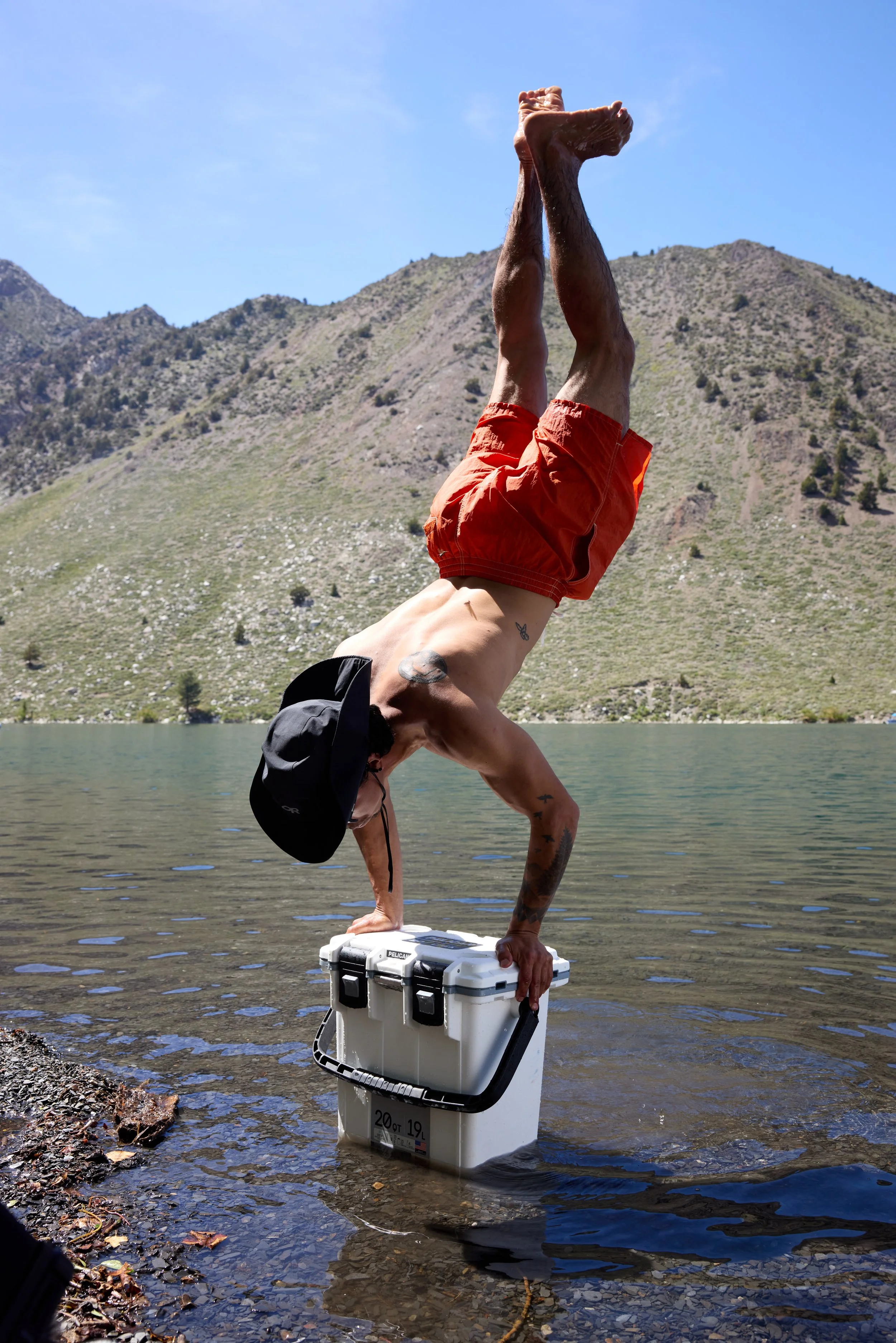 Man performing a handstand on a white cooler by a lake, with mountains and a clear blue sky in the background.