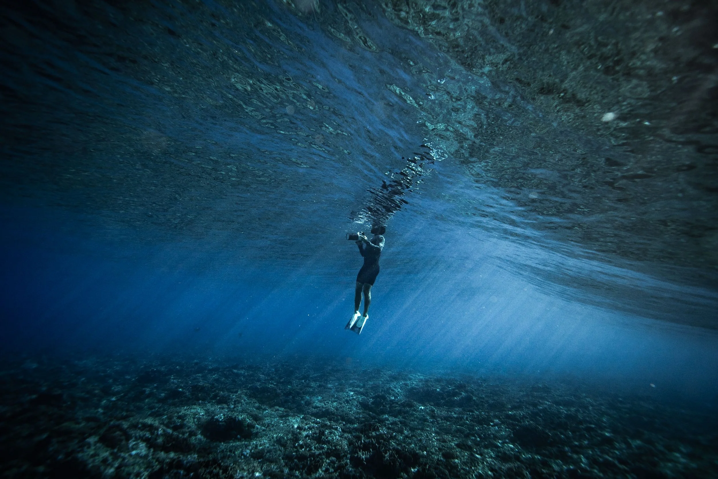 A person swimming underwater in the ocean, holding a device, with sunlight illuminating the water and a rocky ocean floor visible below.
