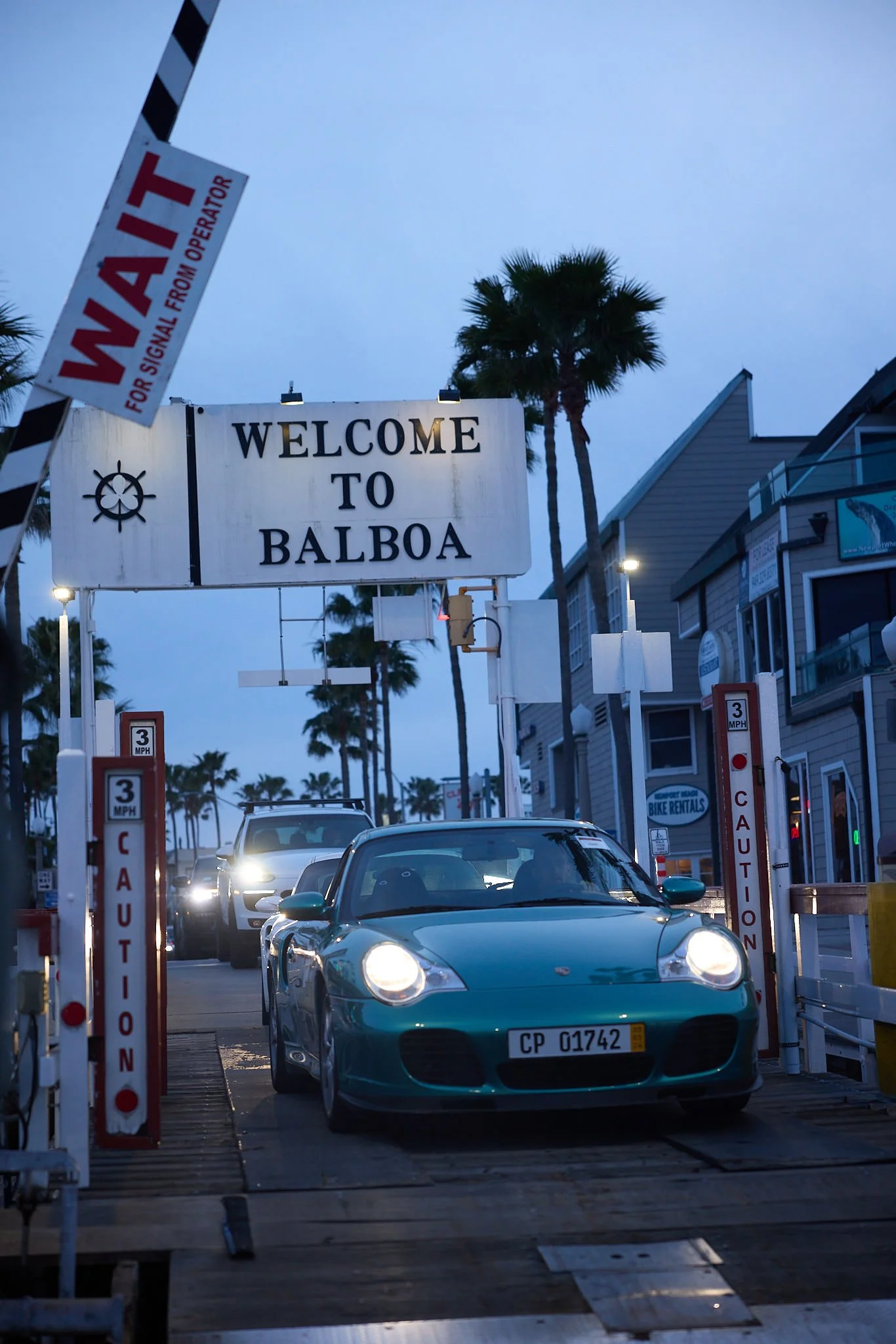 A luxury sports car in a parking lot with a welcome sign for Balboa, palm trees in the background, and a sky transitioning from dusk to evening.
