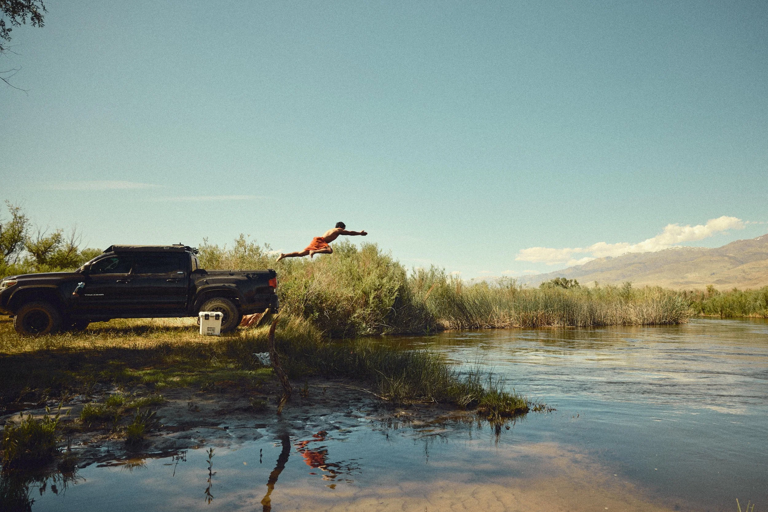 A person in orange shorts is diving from the back of a black pickup truck by a river in a rural area with mountains in the background.