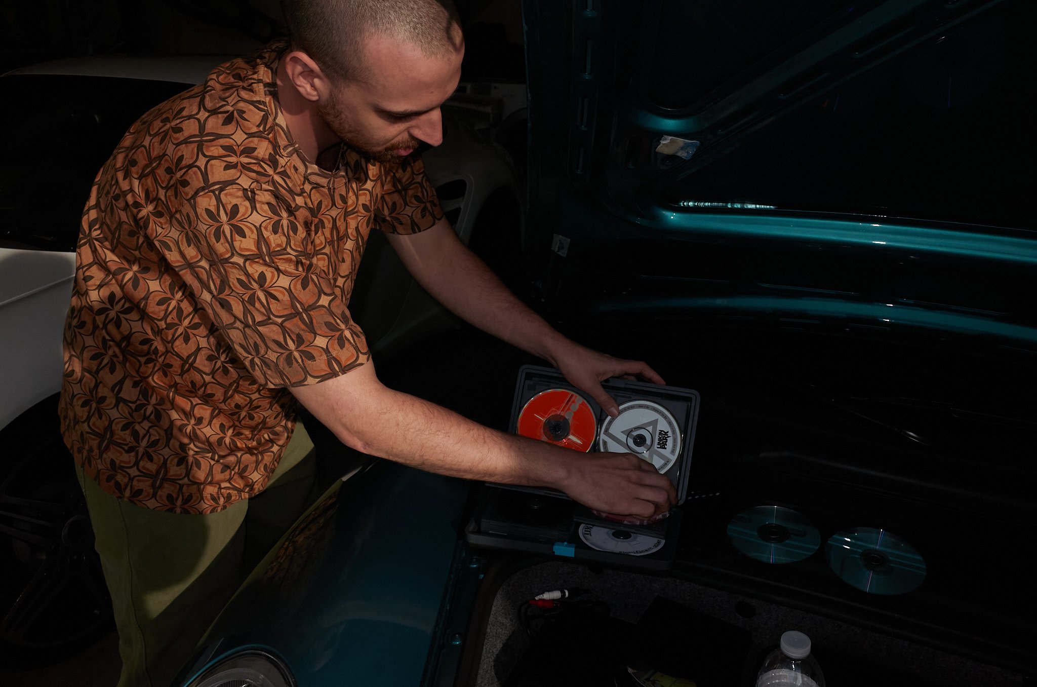 A man wearing a patterned shirt is working on a DJ turntable, adjusting the vinyl record in a dimly lit room with a vehicle in the background.