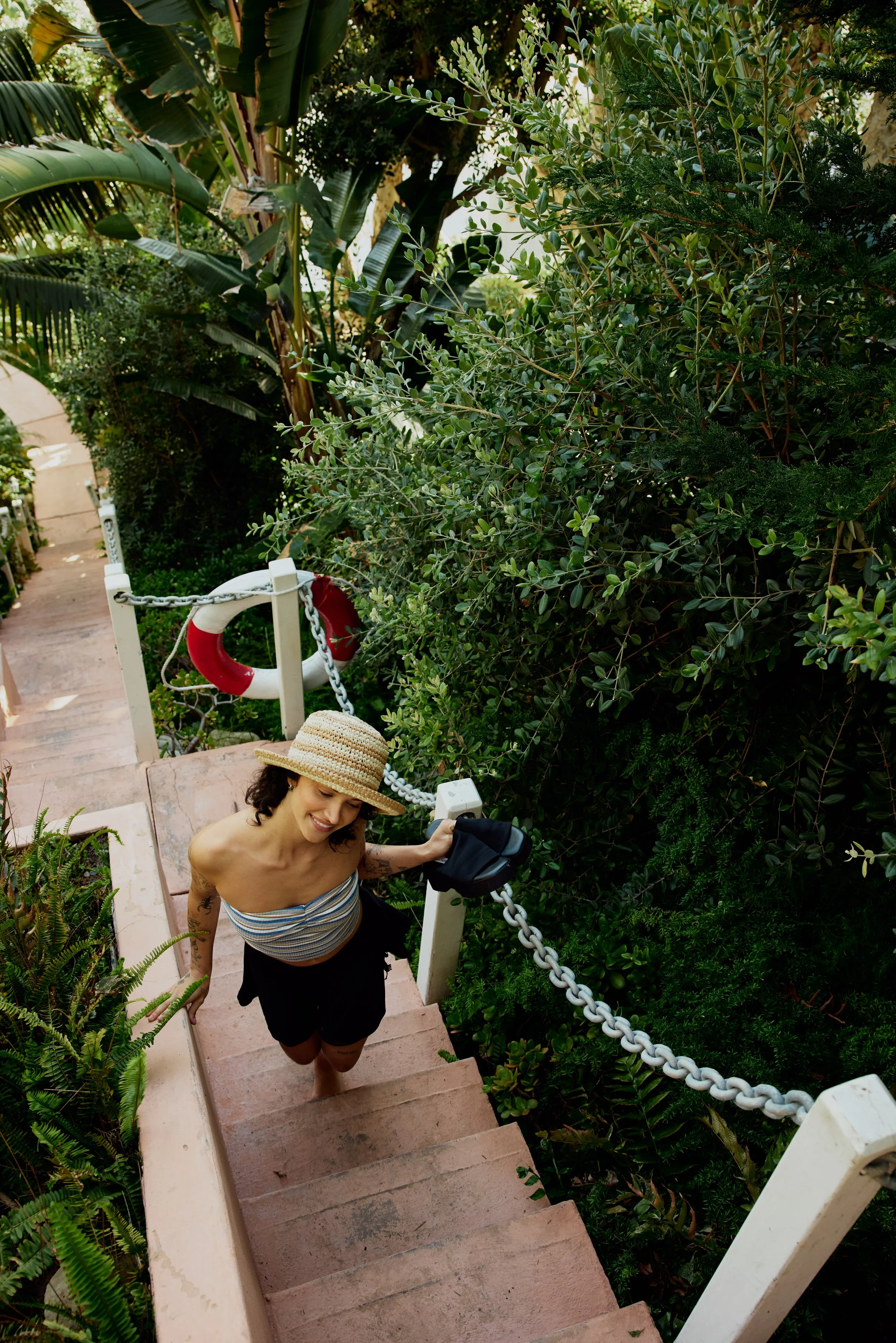 A woman with a straw hat climbing outdoor stairs, surrounded by green plants and bushes, with a body of water or sky visible in the background.