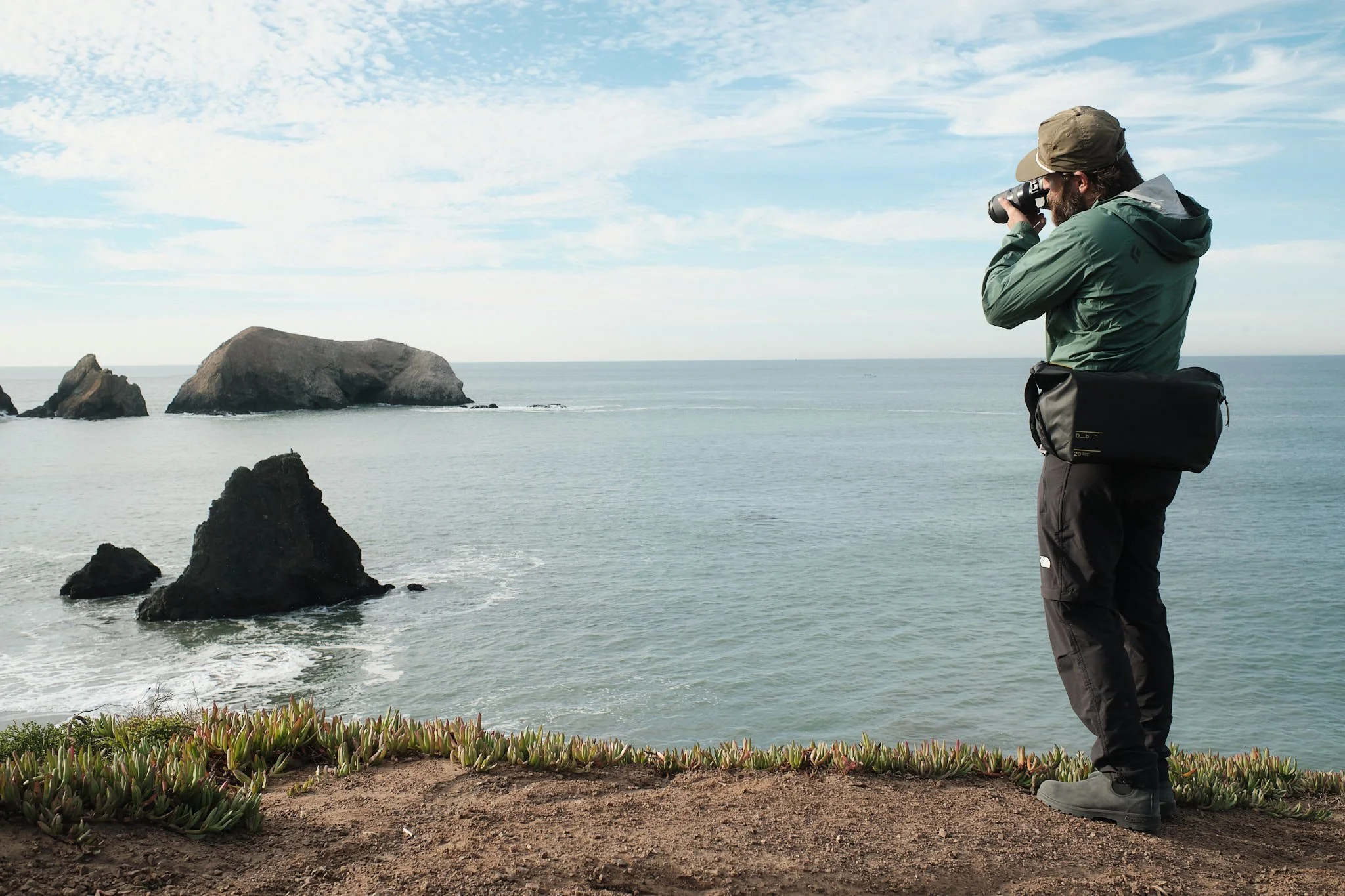 A man with a beard wearing a green jacket, hat, and sunglasses, standing near the edge of the coast, looking through binoculars at the ocean with rocks in the water and a partly cloudy sky.