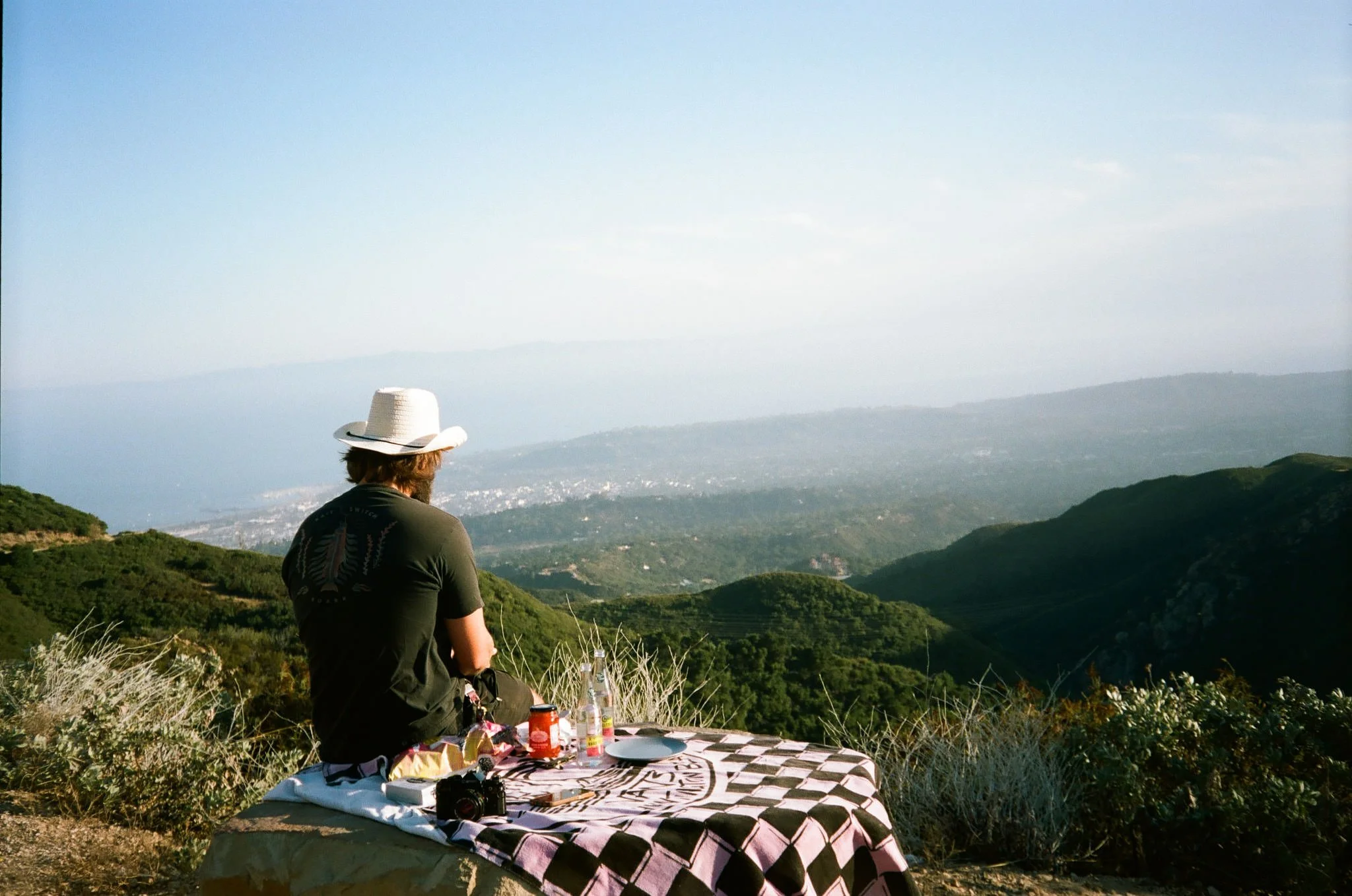 A person with a cowboy hat sitting outdoors on a grassy hill, overlooking a valley with hills and distant mountains, with a table set with food, drinks, and a camera.