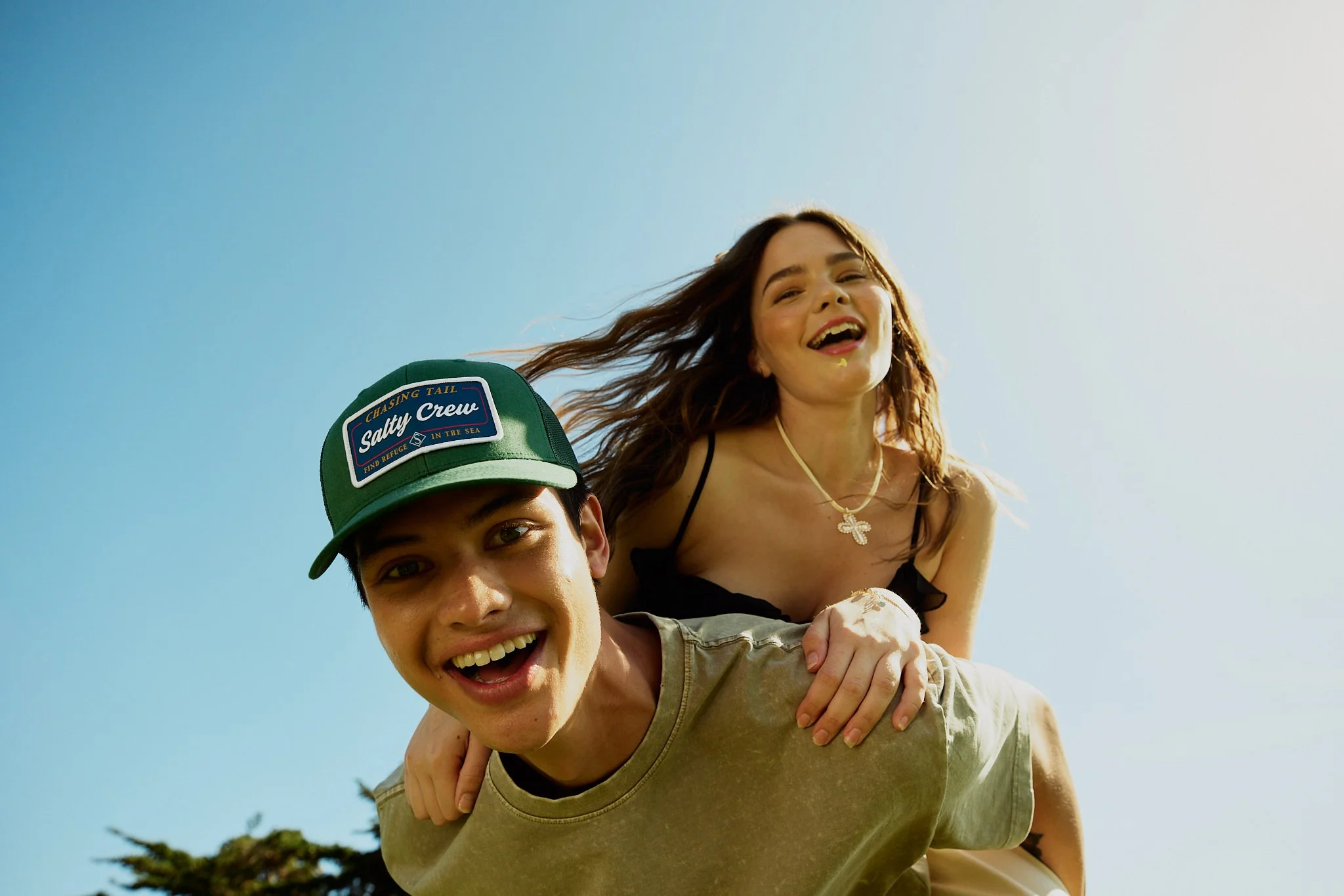 Two young adults, a man and a woman, smiling and enjoying a piggyback ride outdoors on a sunny day, with a clear blue sky in the background.