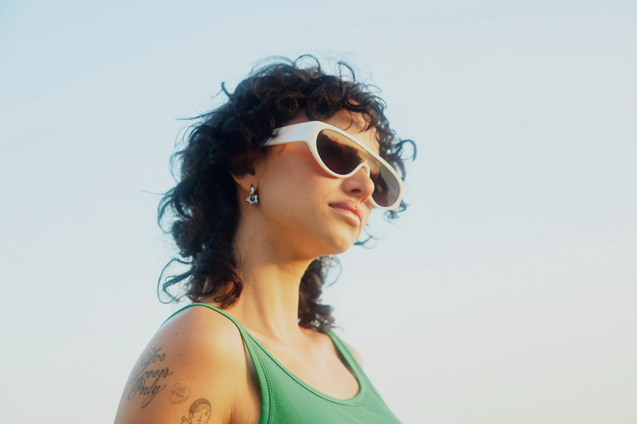 A woman with curly dark hair wearing white sunglasses and star-shaped earrings, wearing a green tank top, looking to the right with a smile, against a clear sky.