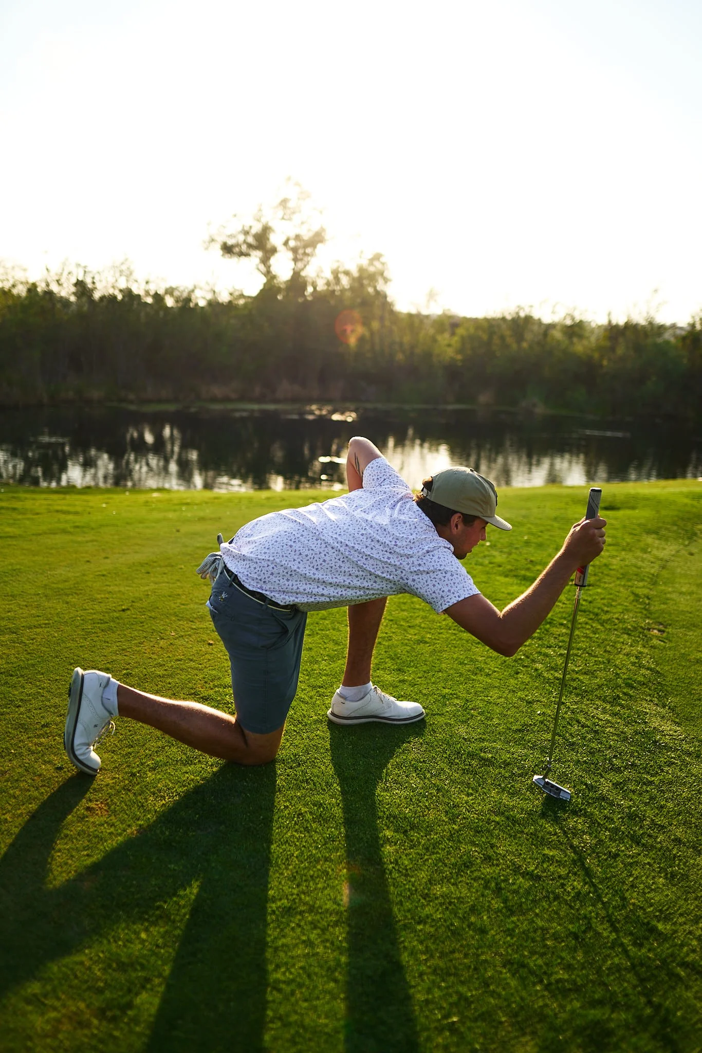 A man in a white shirt, gray shorts, white sneakers, and a gray cap is kneeling on one knee on a grassy area, taking a photo of a golf hole marker with a smartphone as the sun sets behind trees across a small body of water.