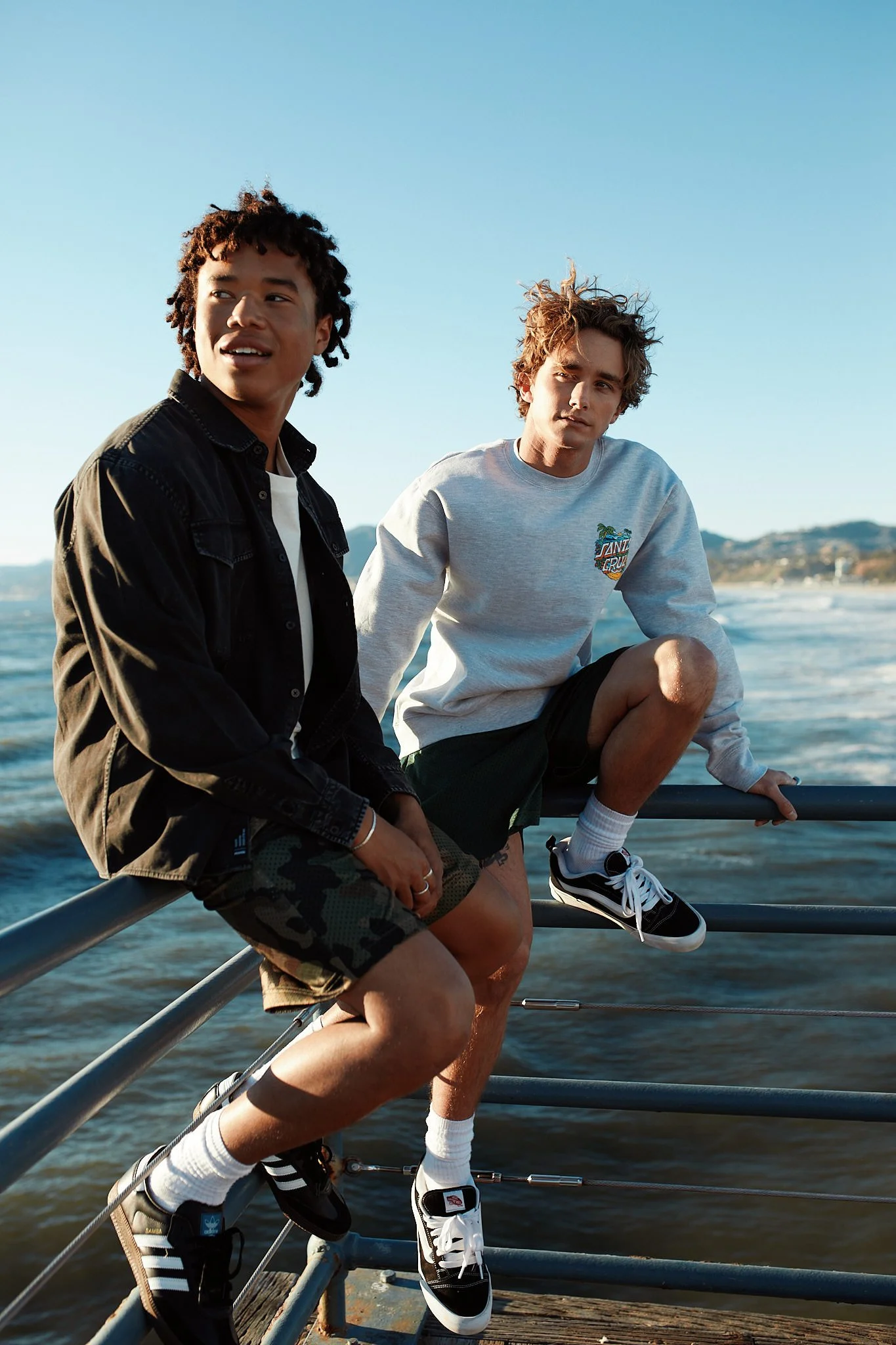 Two young men sitting on a railing by the ocean on a sunny day, with land visible in the background. One is wearing a black jacket and camo shorts, the other a light grey sweatshirt with a graphic, and both are wearing sneakers and socks.