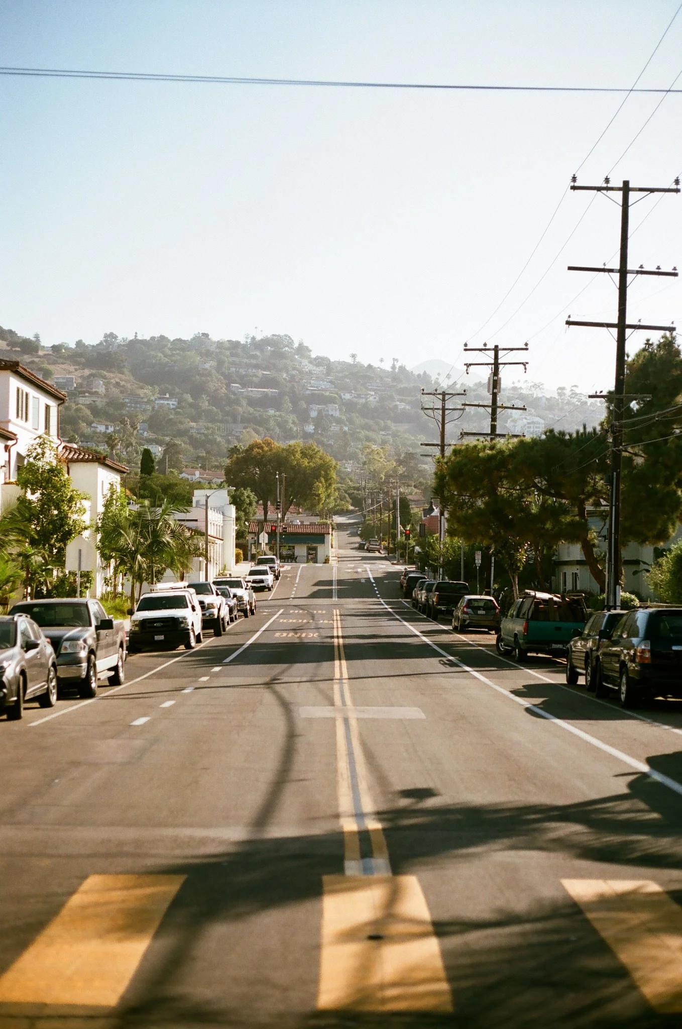 A street scene in a residential area with parked cars lining both sides of the street, trees along the sidewalk, houses in the background, and utility poles with power lines running along the right side of the street. Hills covered with houses are vi