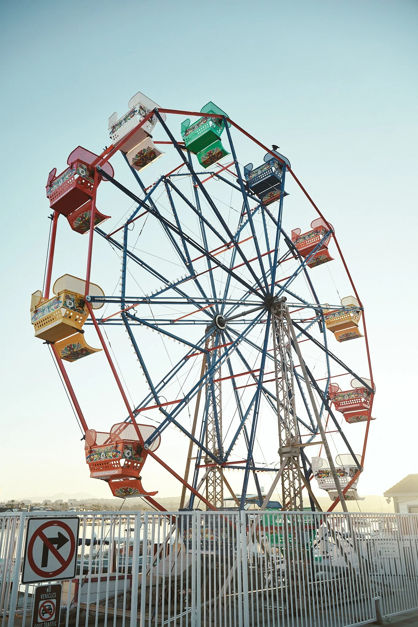An amusement park ferris wheel with colorful gondolas, surrounded by a white fence, with signs indicating no vehicles or traffic allowed, set against a clear sky with distant cityscape.