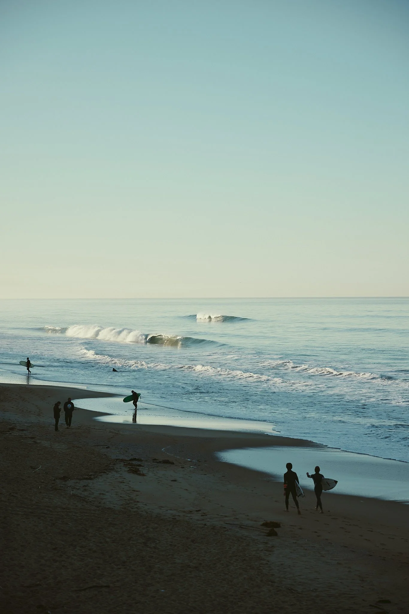 People on a beach during sunset with a surfer carrying a surfboard, some individuals standing, and waves in the ocean.
