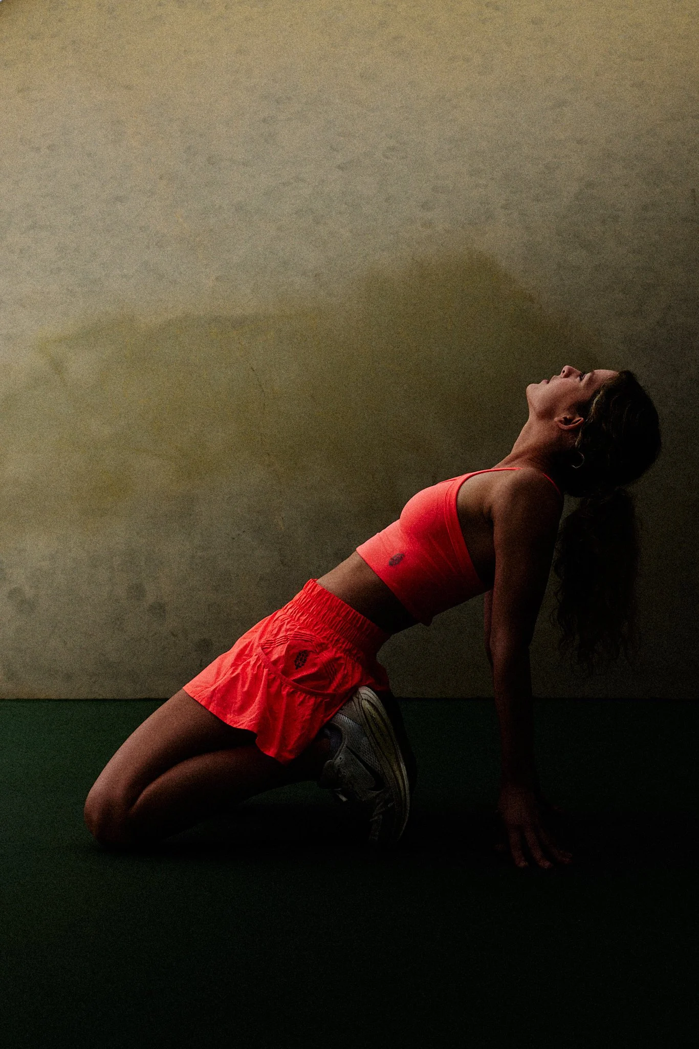 A woman with long dark hair in a ponytail, wearing a bright orange sports bra and matching shorts, is exercising against a plain wall. She has her right knee on the ground, her left leg bent, and her left hand is supporting her body on the floor. She