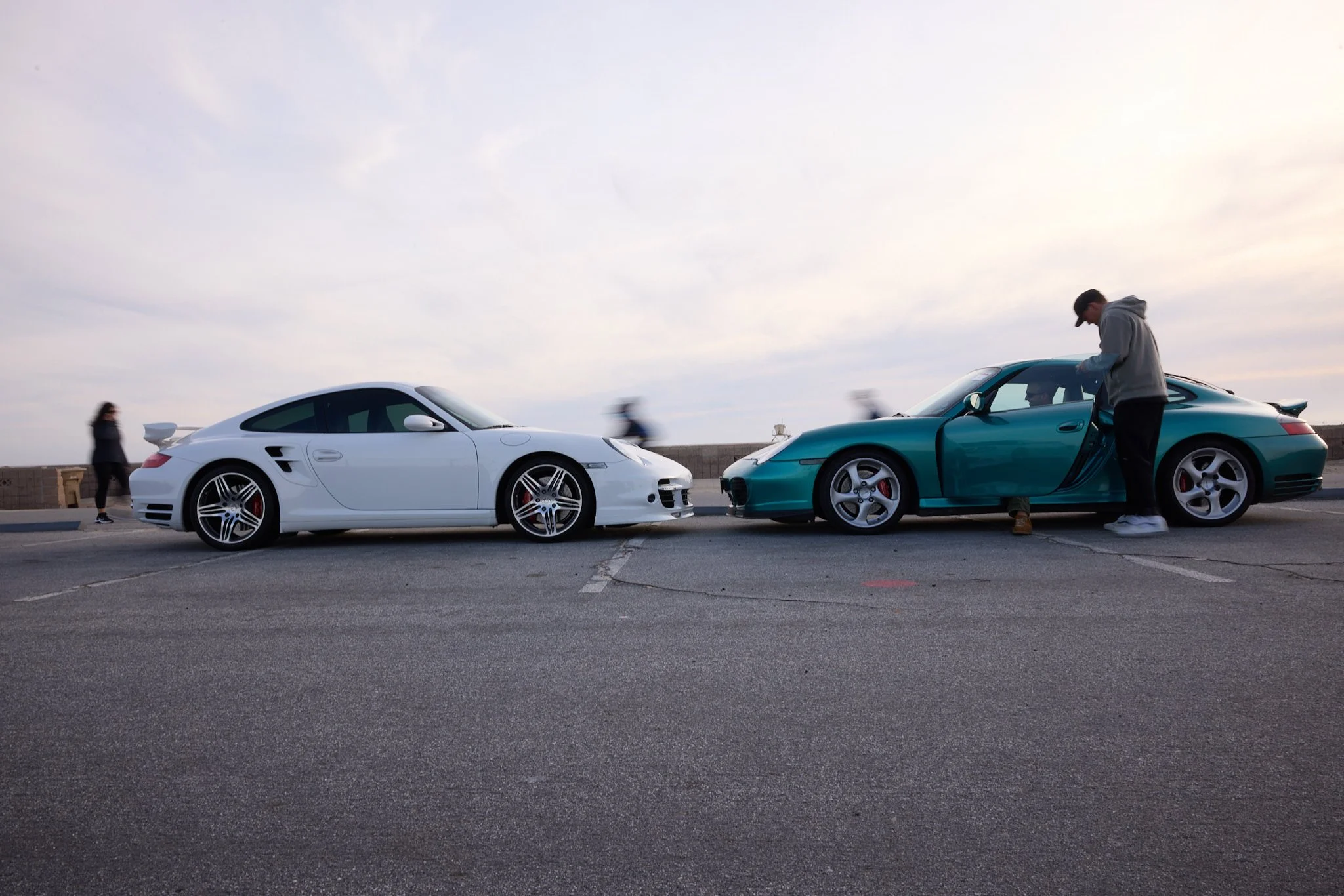 Two sports cars, one white and one blue, parked side by side on a pavement near the water. A man is standing outside the blue car, possibly working on it or inspecting it. The scene is set outdoors with a few people walking in the background and a sk
