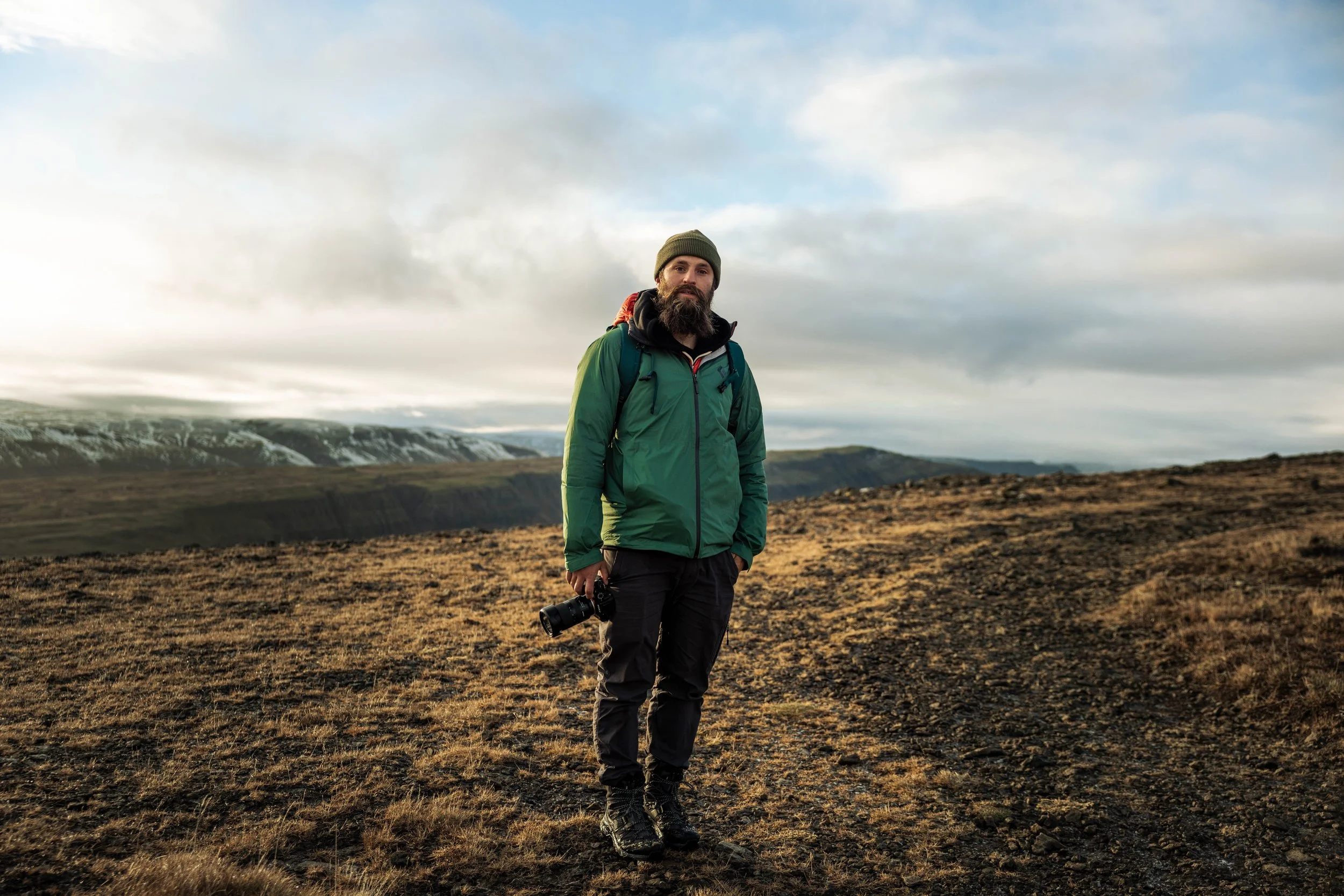 A man with a beard and wearing a green jacket, gray pants, hiking boots, and a beanie, standing outdoors with a camera in his hand, in a vast open landscape with mountains and a cloudy sky in the background.