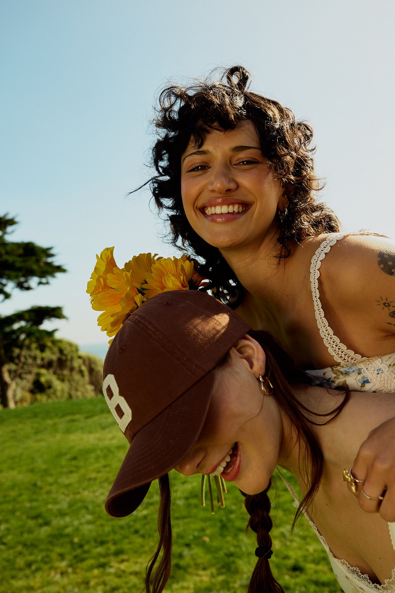 Two women playing with sunflowers outdoors, one wearing a brown cap and the other smiling with curly hair and a lace top, with a green field and trees in the background.