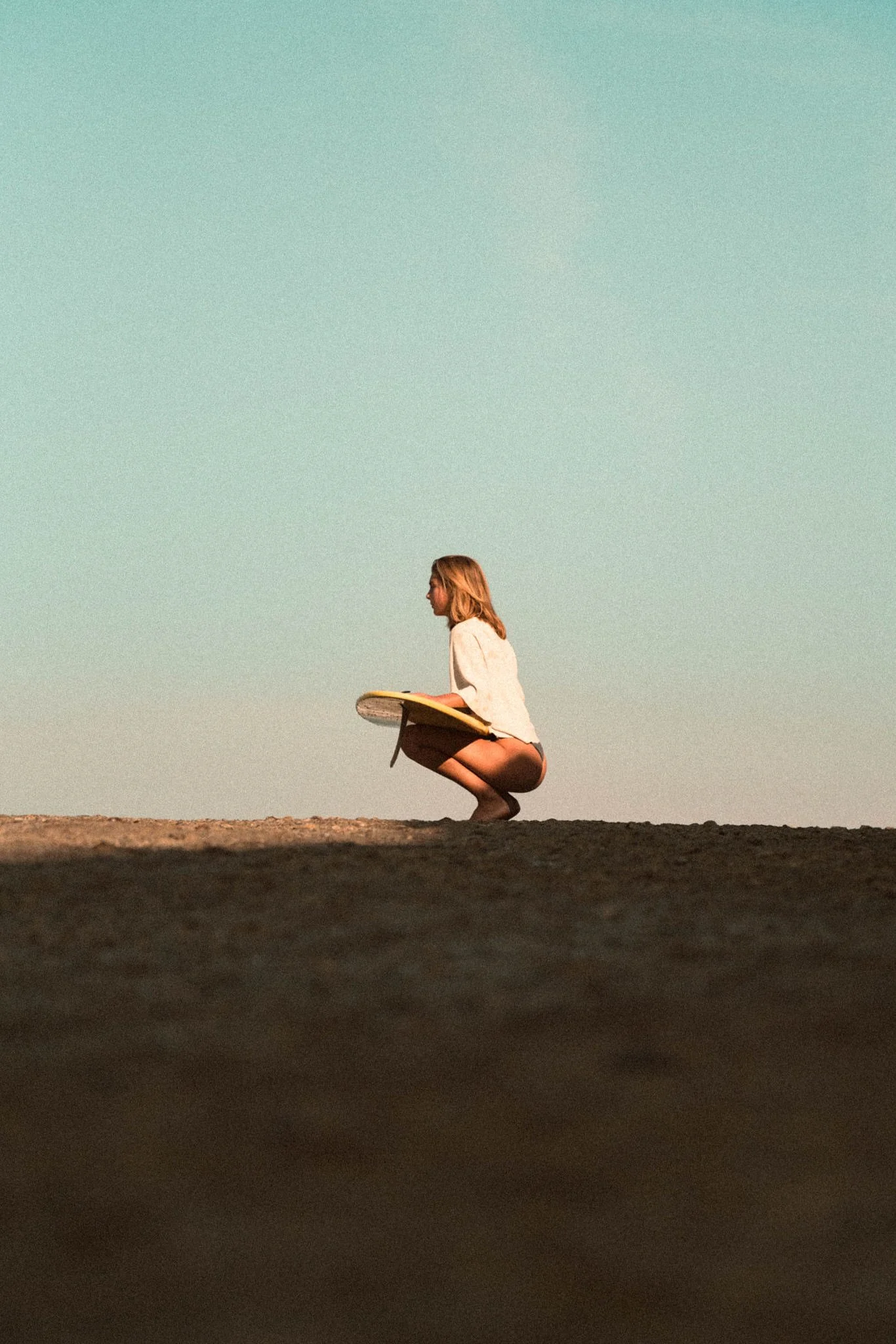A woman squatting on a beach holding a surfboard, wearing a white long-sleeve shirt and shorts, with a clear sky in the background.