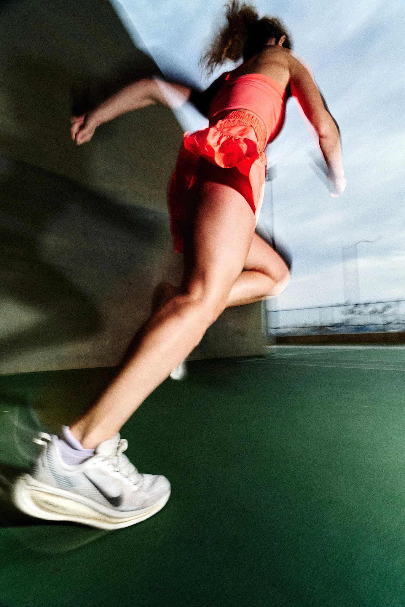 A person running on a track wearing athletic shorts, a tank top, white sneakers, and a wristwatch, with a blue sky background.