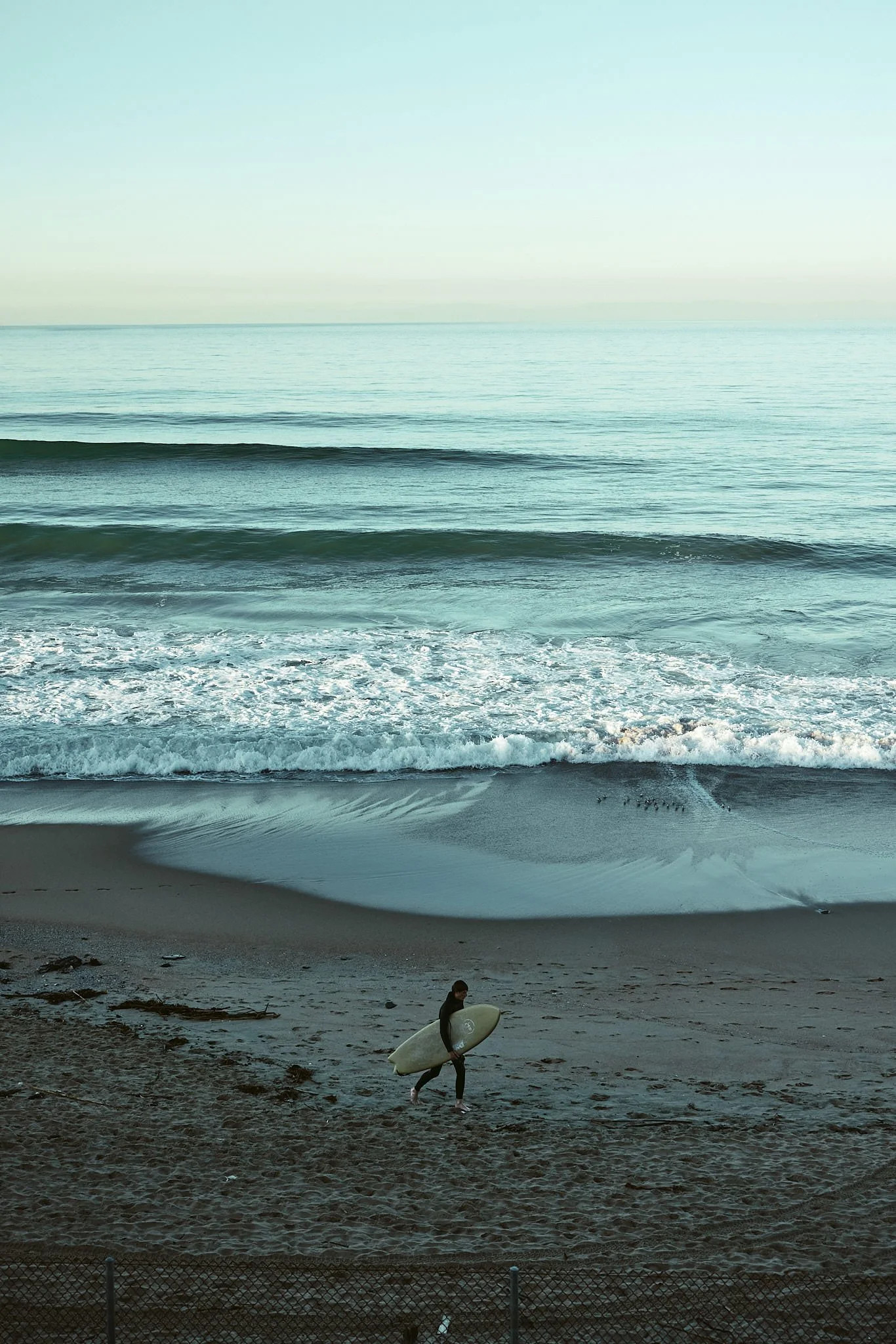A surfer walking on the sandy beach holding a surfboard, with the ocean waves and horizon in the background.