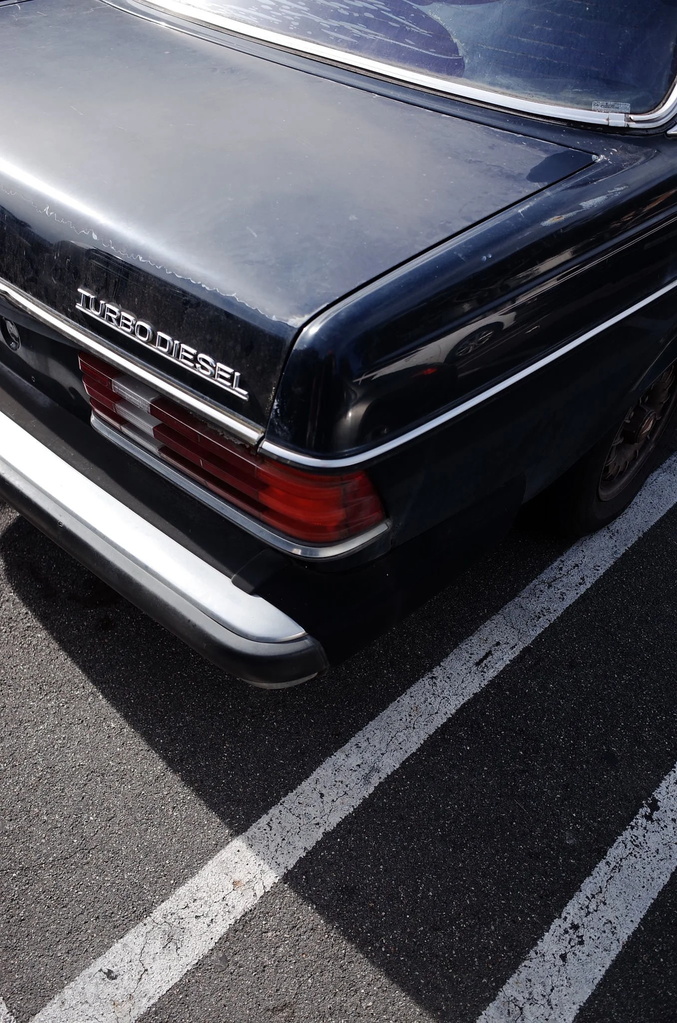 Rear view of a black vintage car with a 'Turbo Diesel' badge, parked in a parking lot with painted white lines, and a shadow cast on the ground.