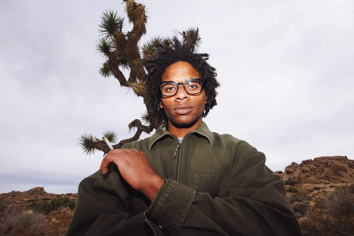 A man with glasses and dreadlocks in a desert landscape with rocky hills and a twisted tree, wearing a green jacket and holding a cigarette.