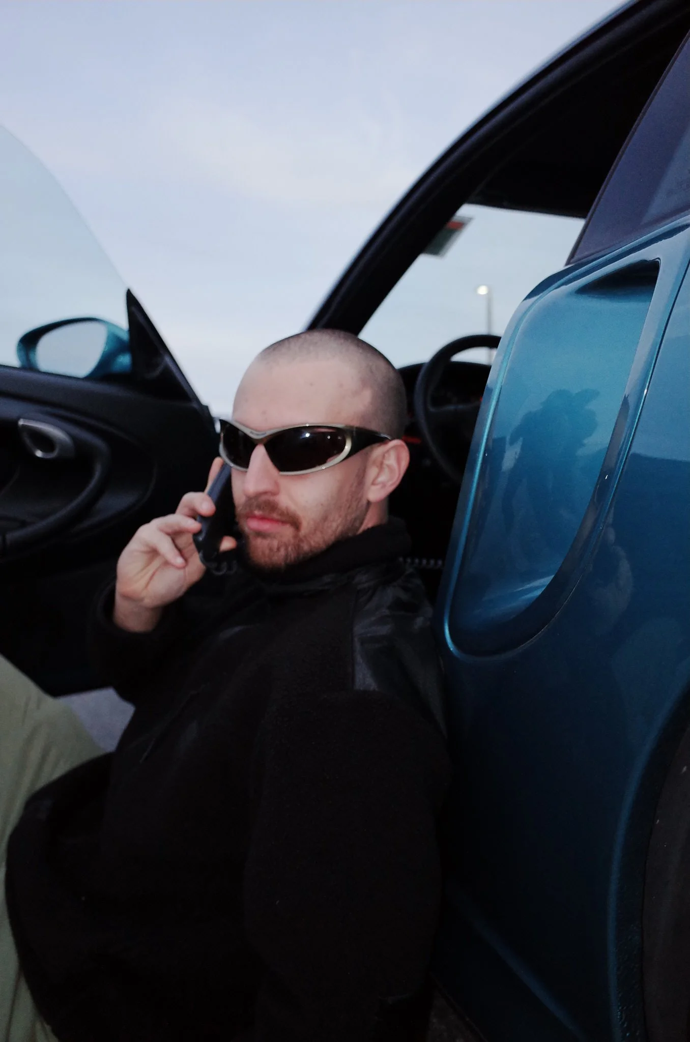 A man with a shaved head and sunglasses sitting in a blue car, talking on a cell phone.