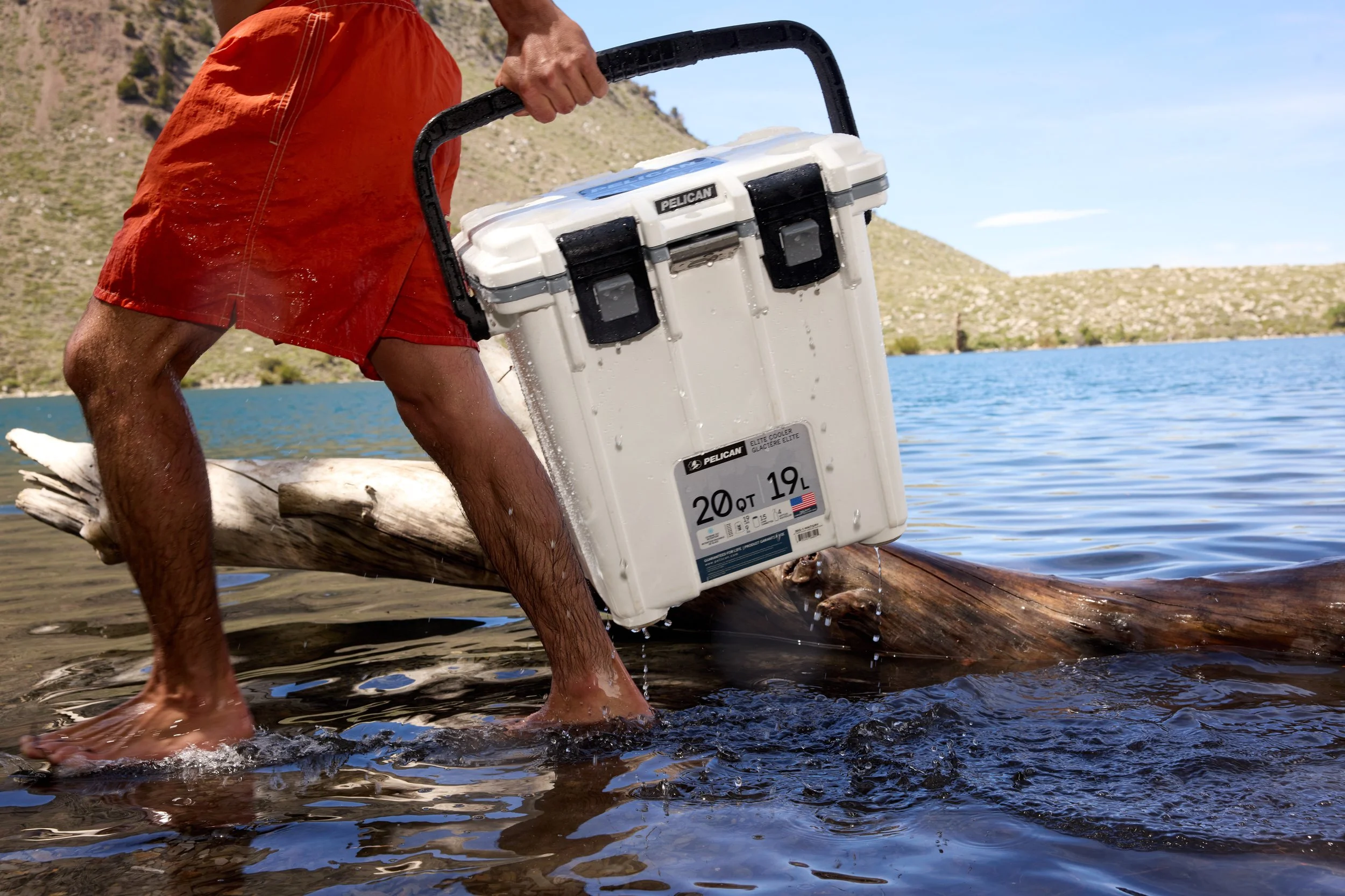 A man in red shorts holding a white Pelican cooler over water, standing on a log at the edge of a lake with hills in the background.