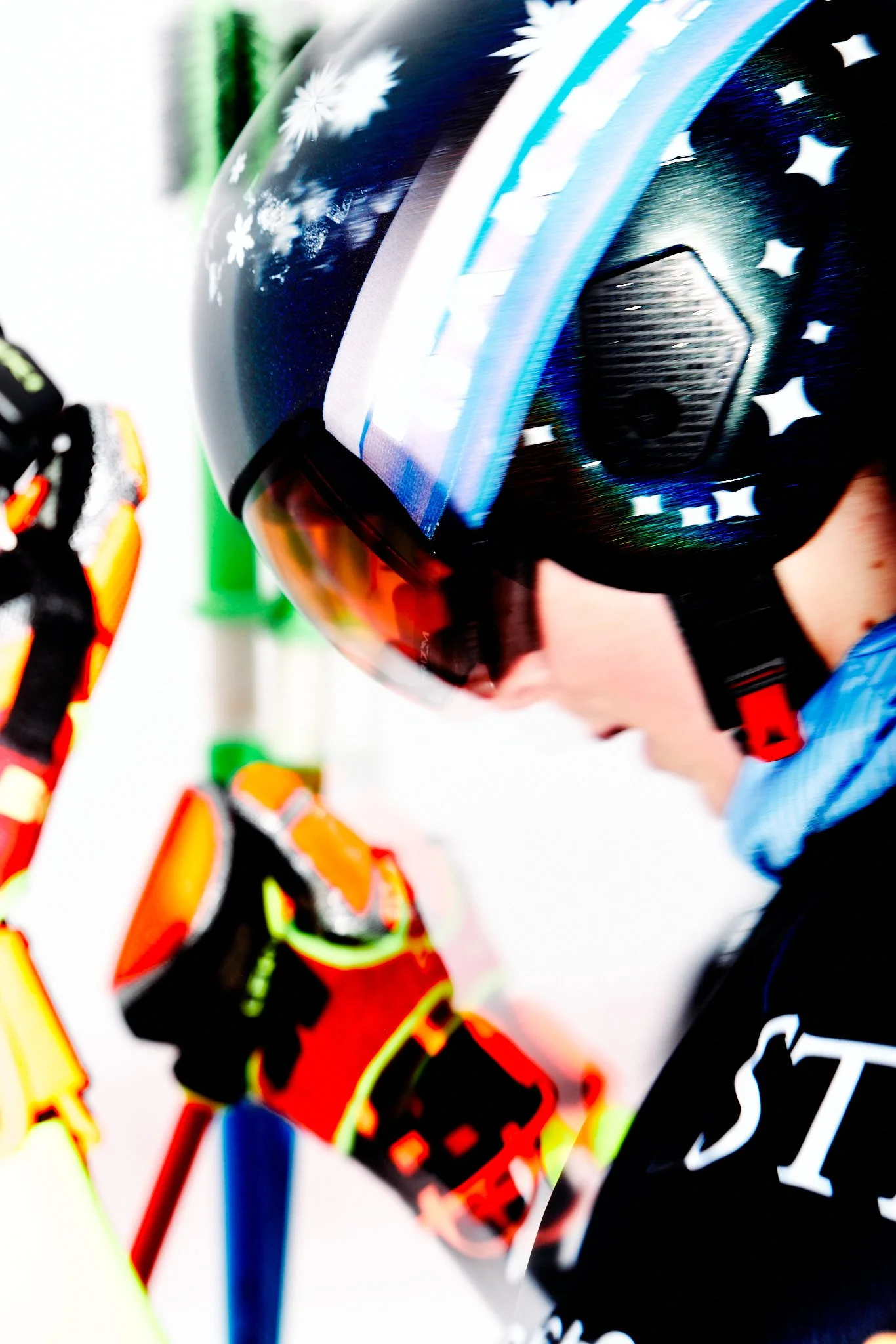 Close-up of a person wearing a black helmet with white snowflake patterns and a reflective visor, with another person in bright orange and yellow gear visible in the background.