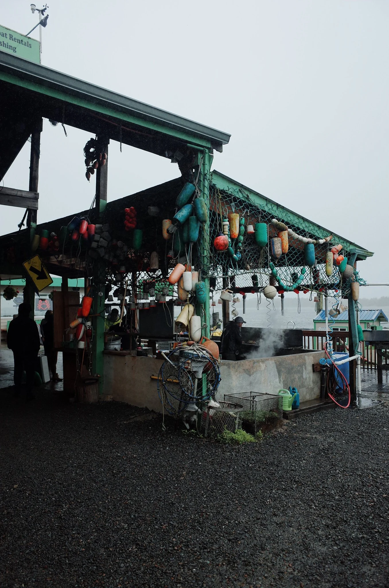 Overcast scene of a fishing dock with various fishing buoys hanging from the roof structure, a barbecue grill with smoke, and some people in the background near water.