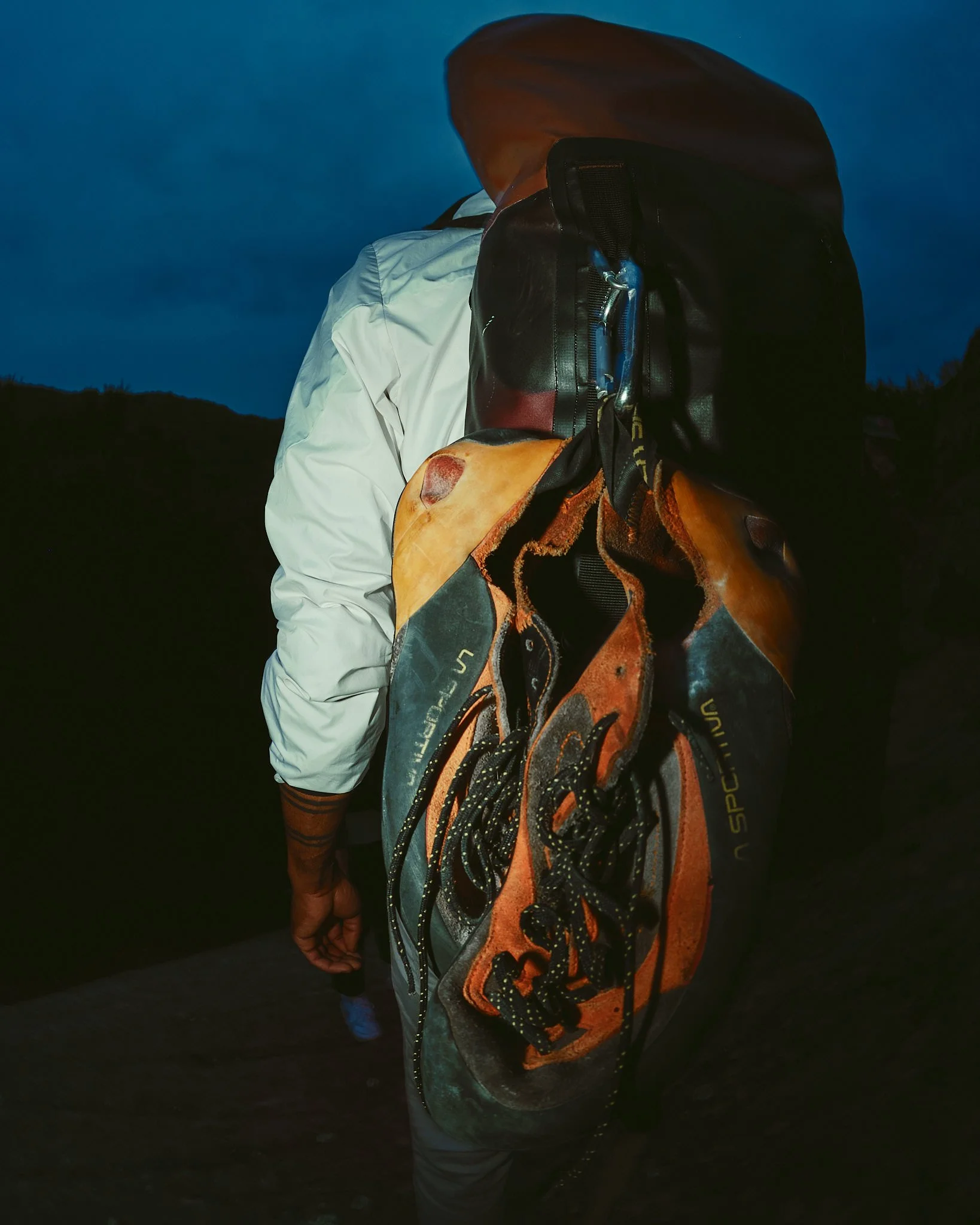 Person wearing a white jacket and a brown hooded jacket carrying an orange and gray backpack, standing outdoors during dusk or night.