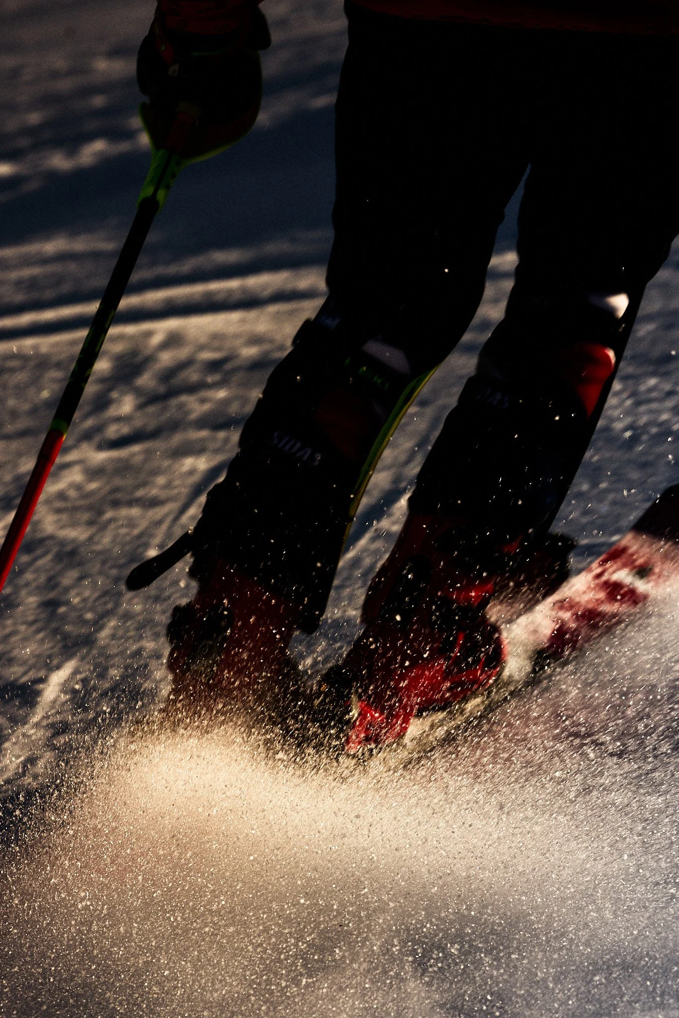 Close-up of a skier's feet and skis moving quickly through snow, kicking up a spray of snow behind.