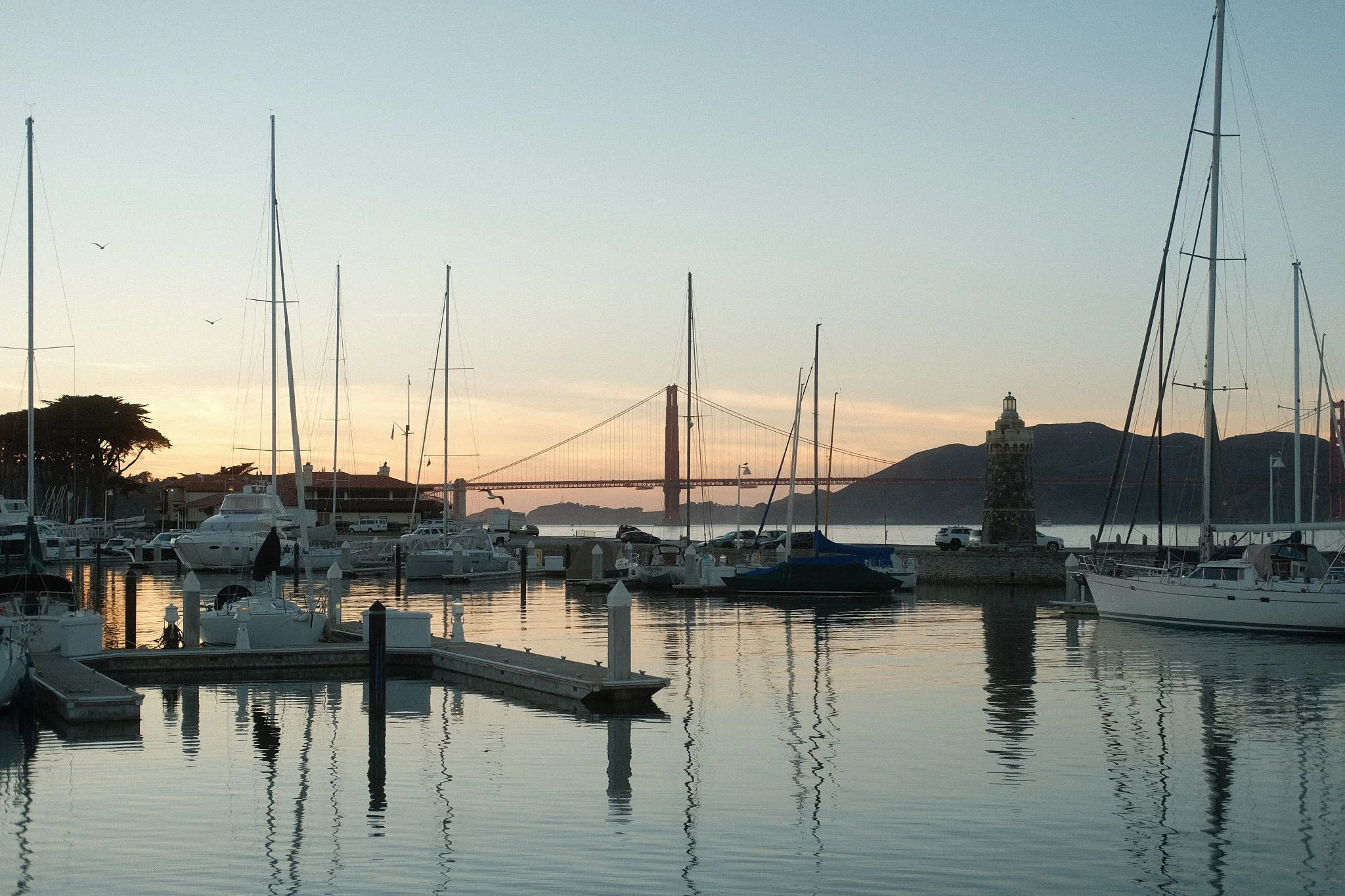 A marina at sunset with boats docked, the Golden Gate Bridge visible in the background, and a lighthouse on the right on a rocky pier.