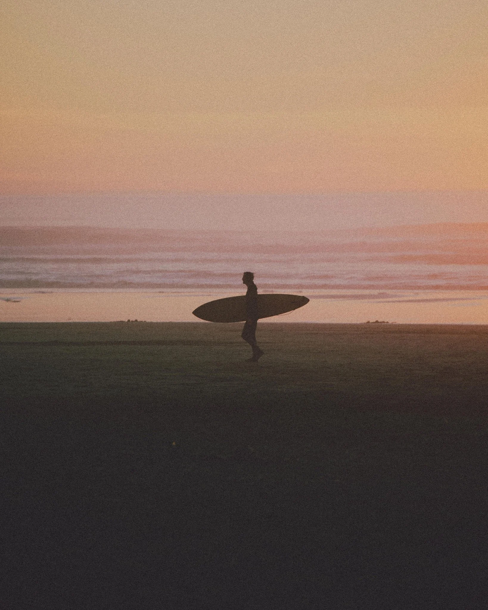 Silhouette of a person holding a surfboard, standing on a beach at sunset with a colorful sky and calm water.