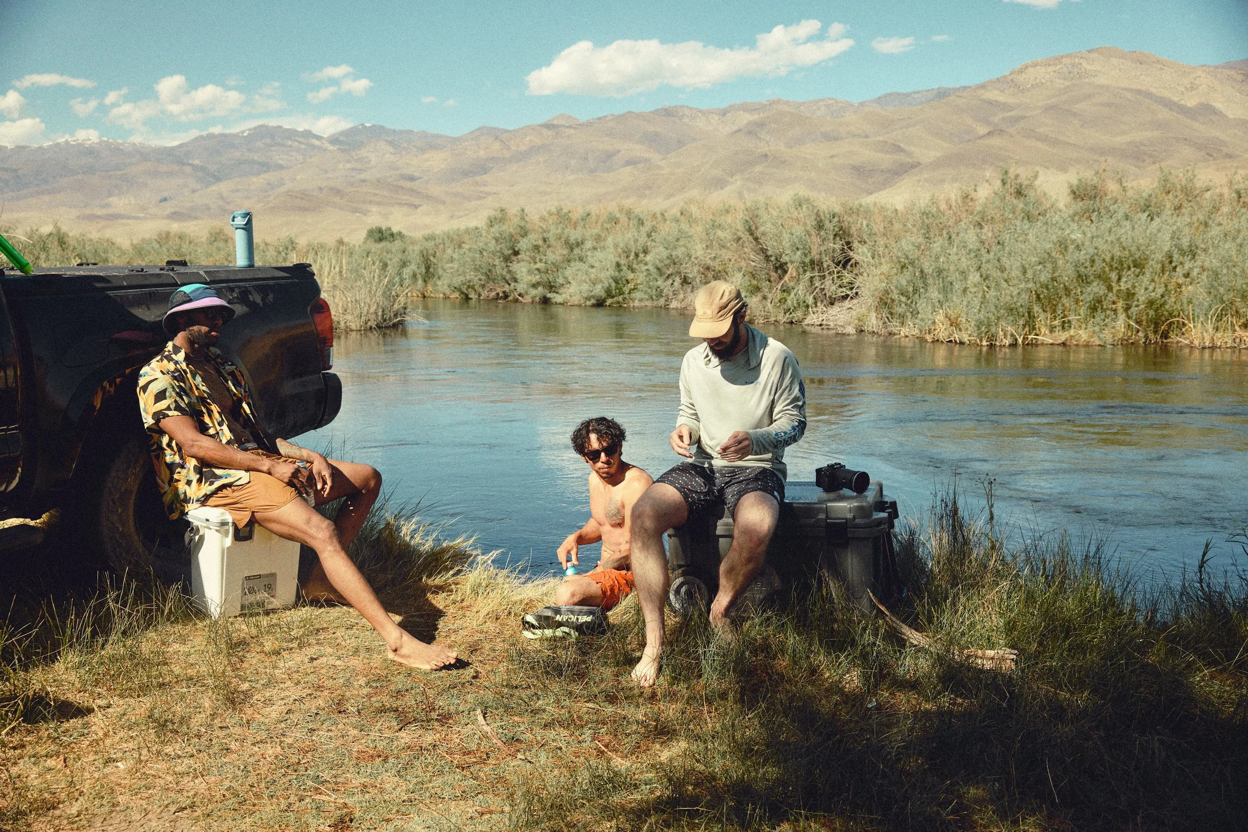 Group of people fishing by a river in a scenic outdoor setting with mountains in the background.