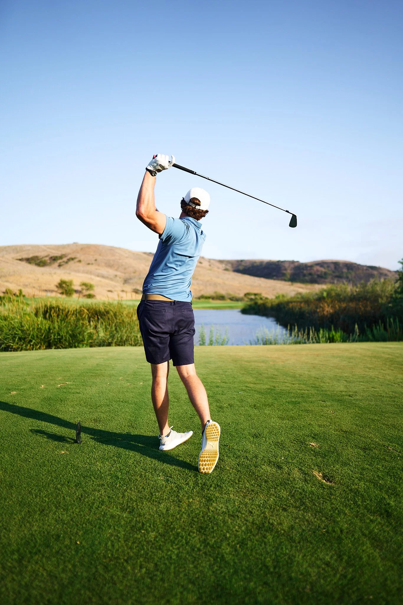 A man in a light blue polo shirt, black shorts, and white golf shoes is swinging a golf club on a golf course near a body of water with rolling hills in the background.