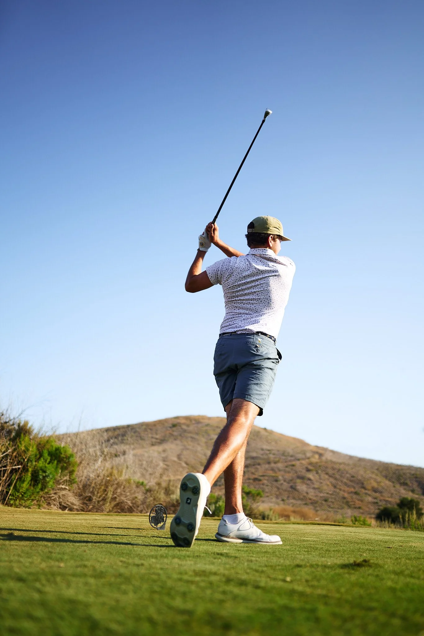 A man in a white patterned shirt, gray shorts, and white shoes playing golf, swinging his golf club on a green golf course with a hill and clear sky in the background.