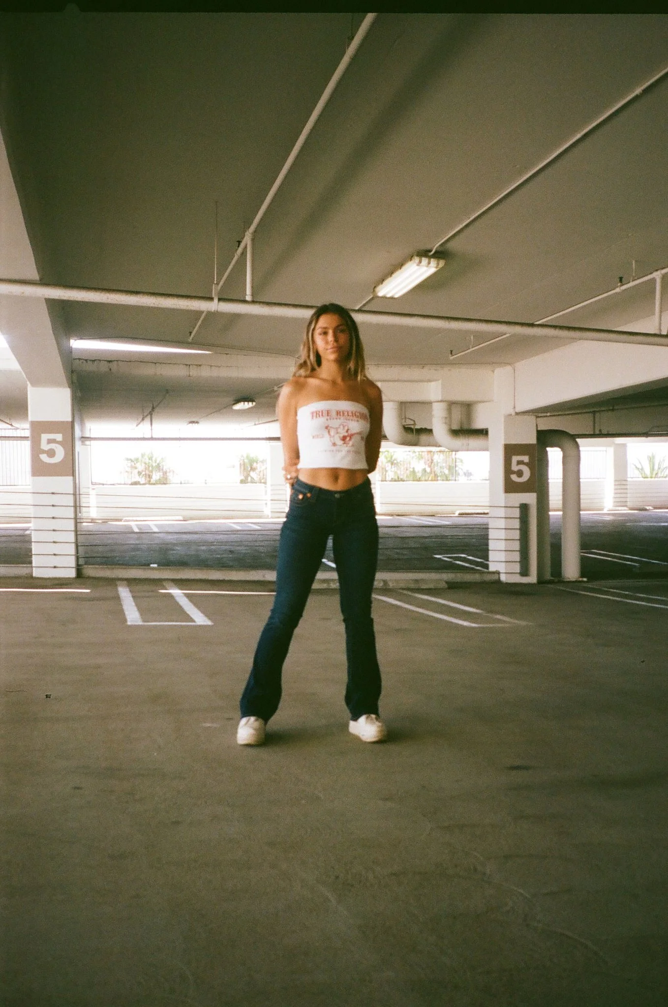 A woman standing in an empty parking garage, wearing a white tube top, dark jeans, and white shoes, with her hands behind her back.