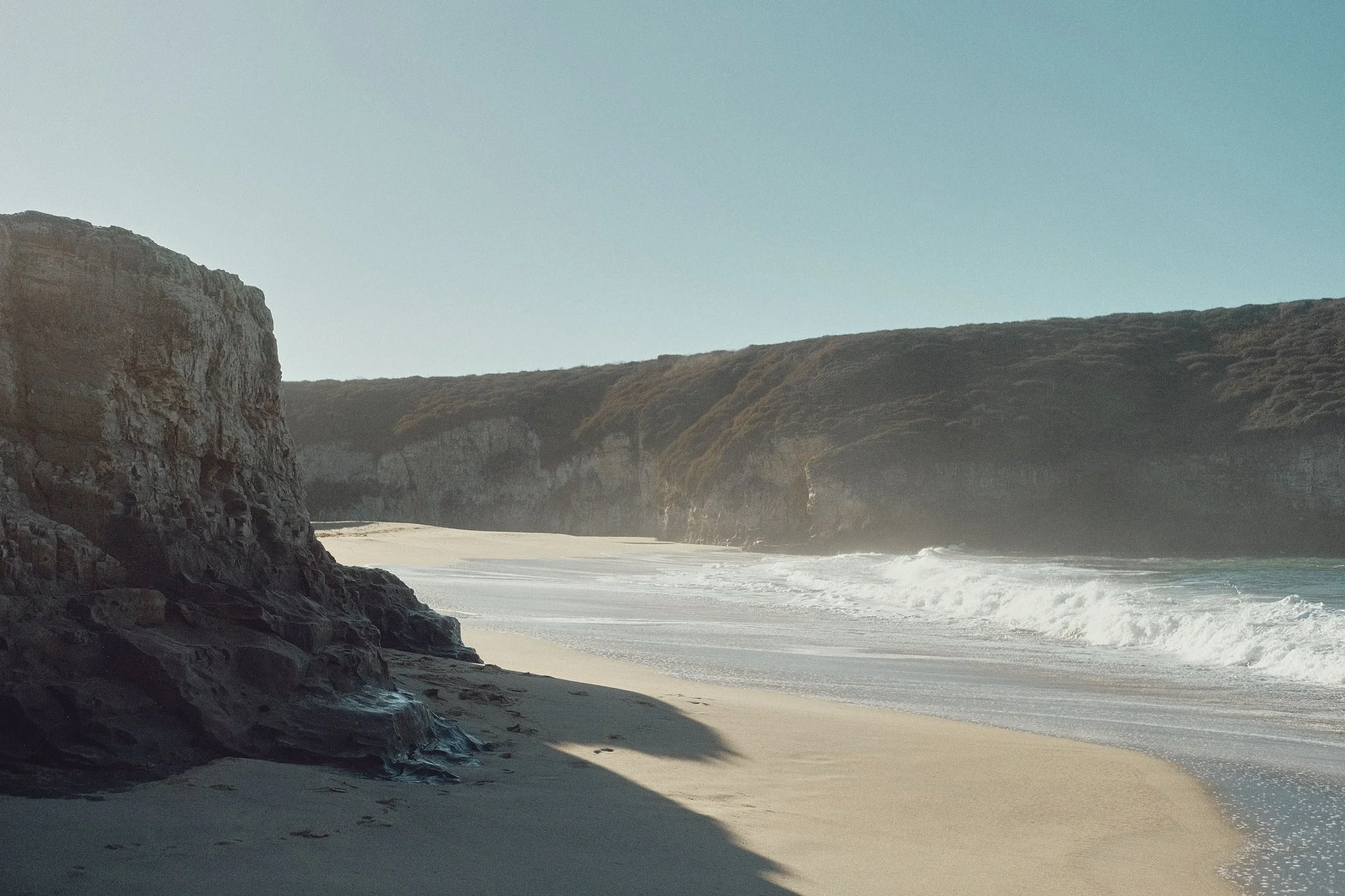 A rocky beach with tall cliffs and waves crashing onto the sandy shore.