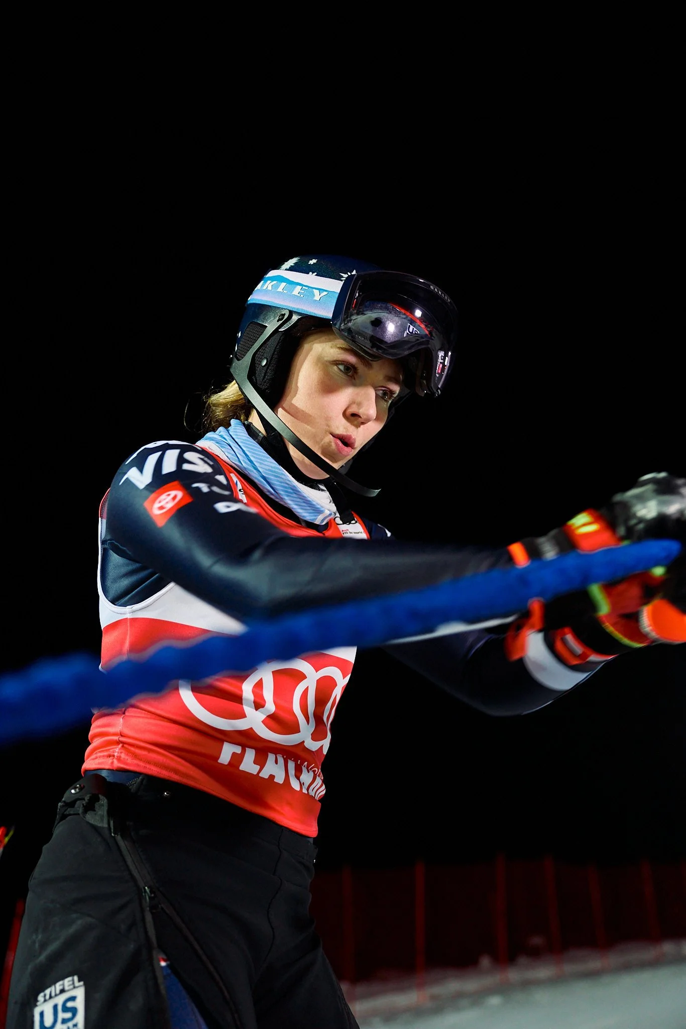 Young female skier with helmet and goggles preparing for a ski run during night or indoor event, wearing a USA team uniform with logos.