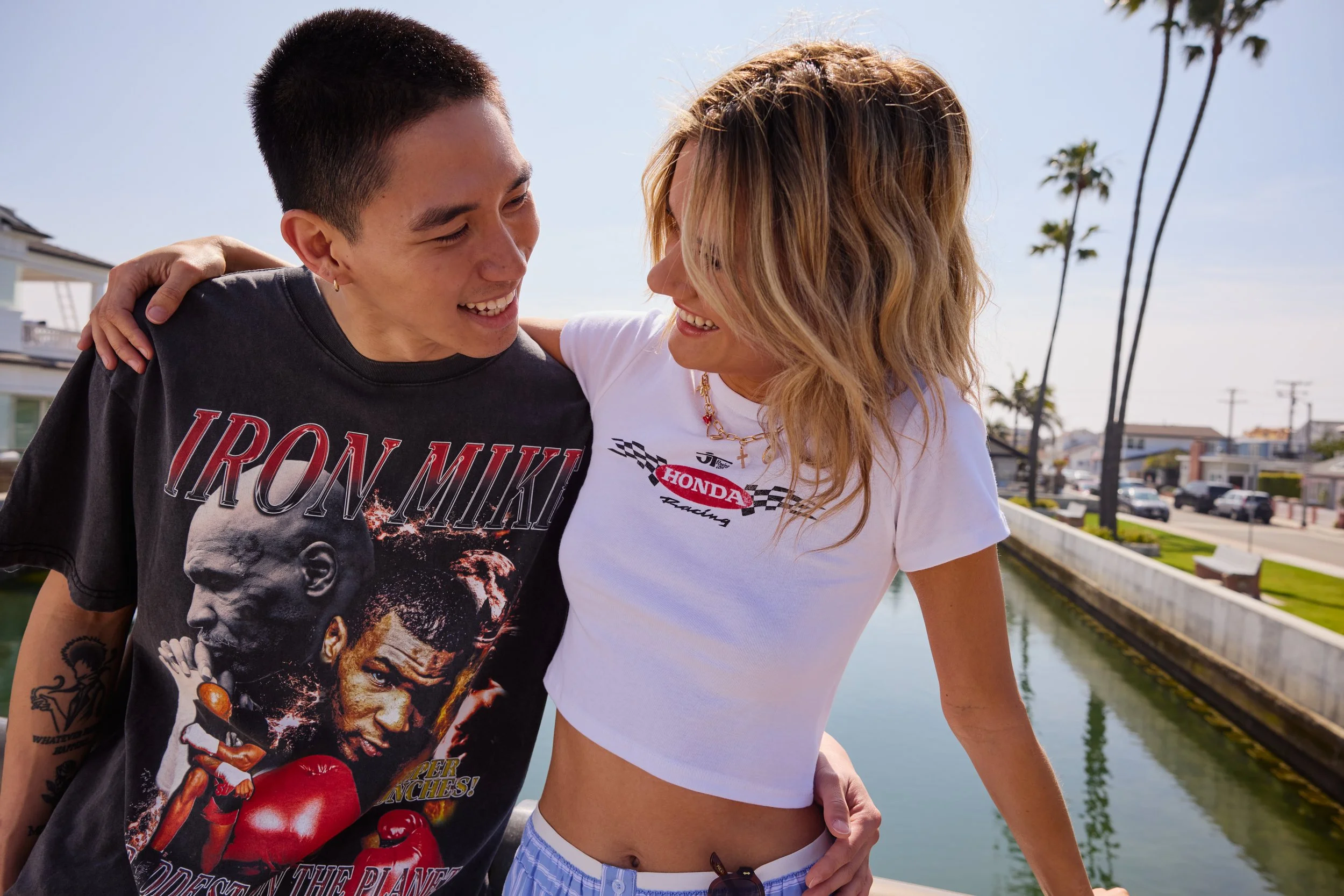 Two people, a man and a woman, smiling and looking at each other outdoors with palm trees and a canal in the background.