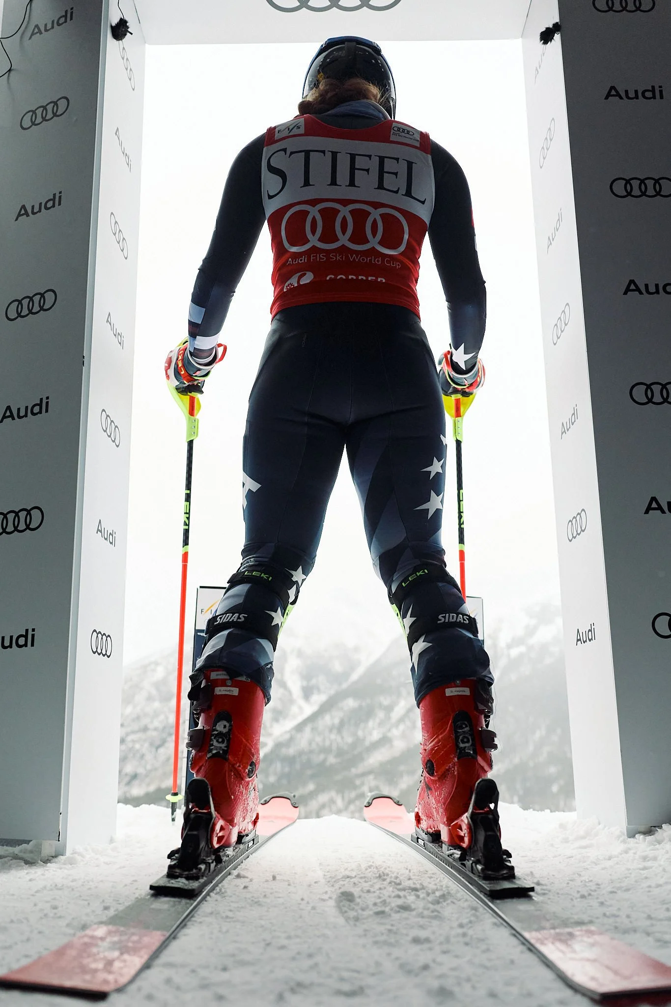 A skier preparing to race on snow, standing at the starting gate, wearing a helmet, goggles, ski suit, and holding ski poles, with snow-covered mountains in the background.