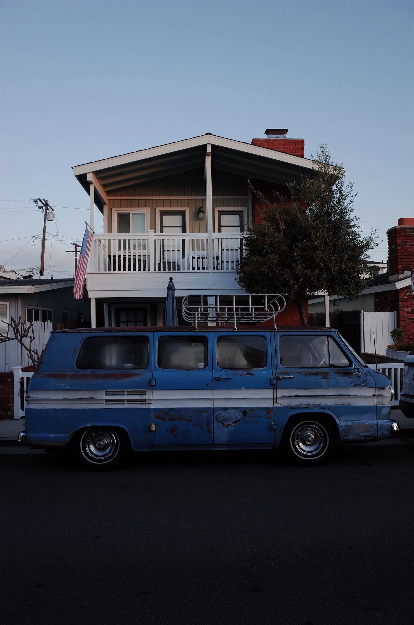 An old, rusty blue van parked in front of a two-story house with a balcony and a tree. The house has a brick chimney and a flag hanging from the balcony.