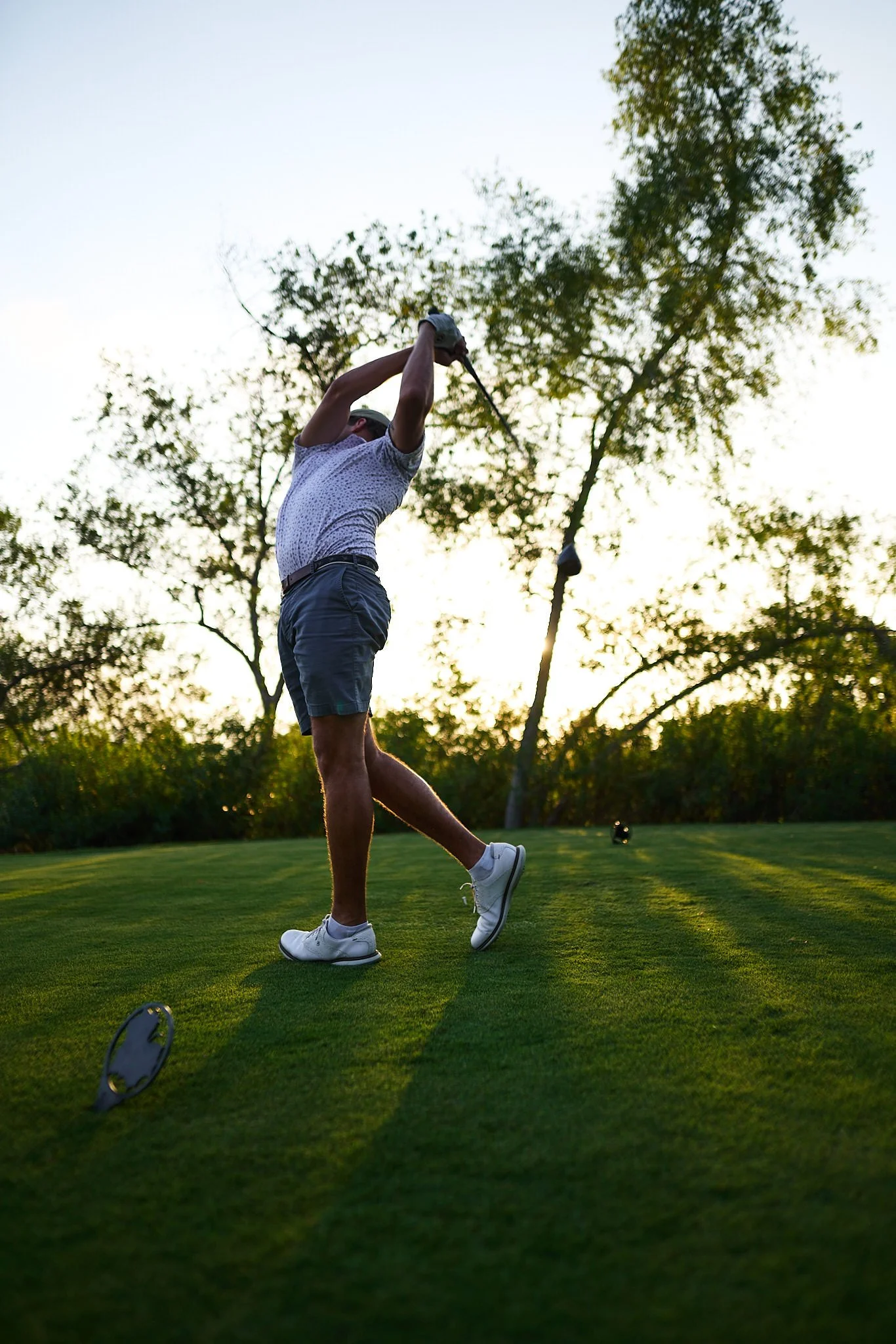 A man swinging a golf club on a golf course during sunset. The man is wearing a white patterned shirt, gray shorts, and white golf shoes. A golf club is on the ground nearby, and trees are in the background.