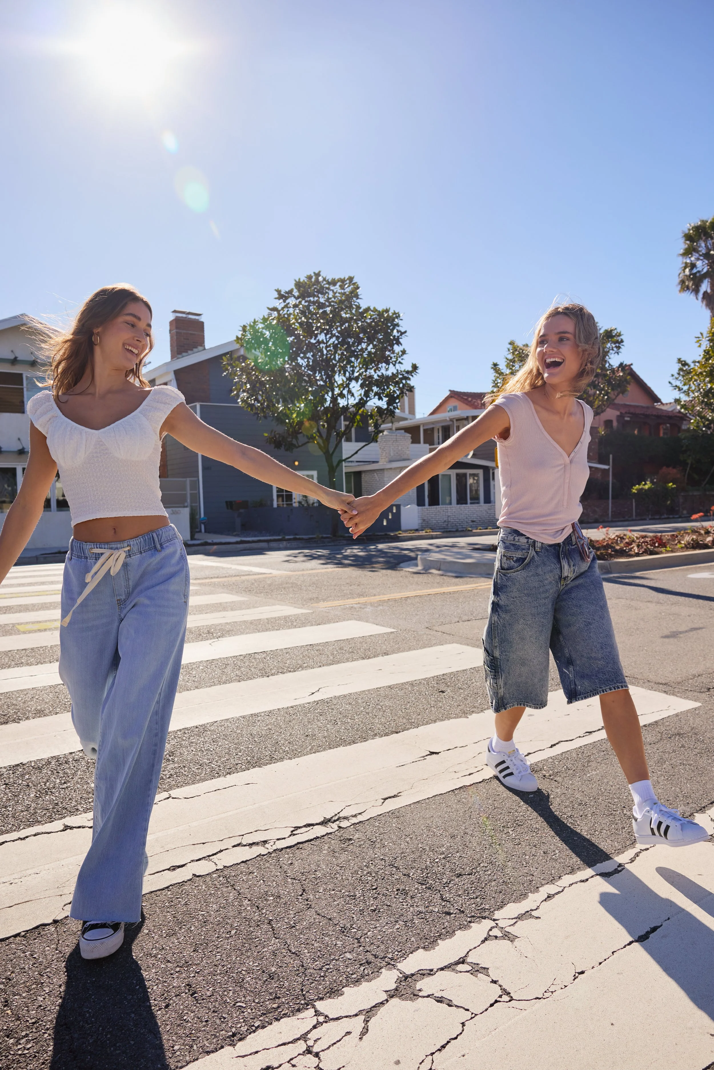 Two young women holding hands and laughing while crossing the street on a sunny day in a residential neighborhood.