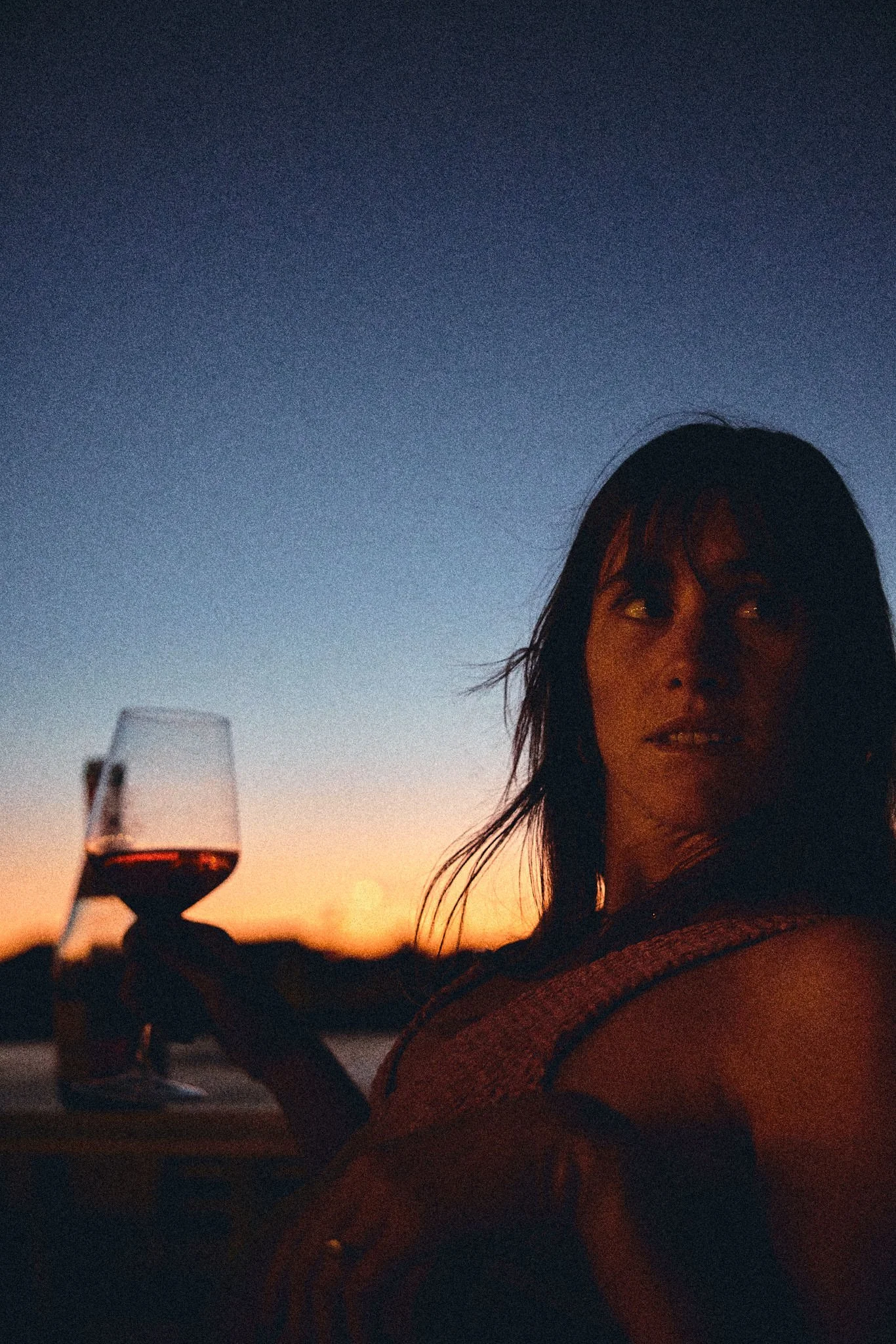 A woman holding a glass of red wine during a sunset or dusk, with a darkening sky in the background.