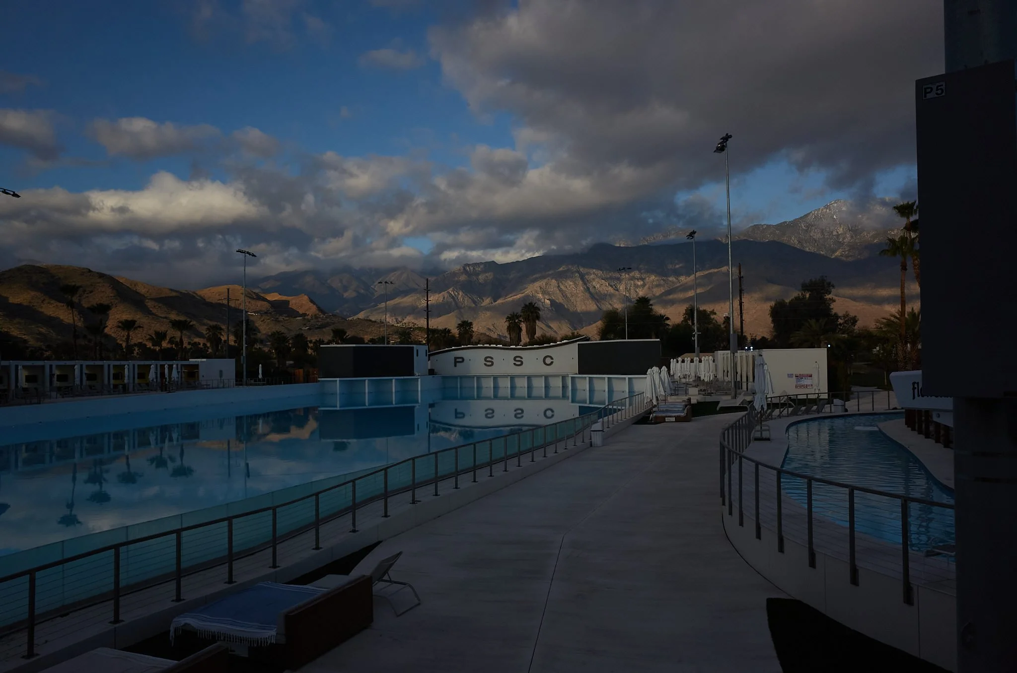 An empty outdoor swimming pool area with lounge chairs, umbrellas, and a view of mountains in the background under partly cloudy skies.