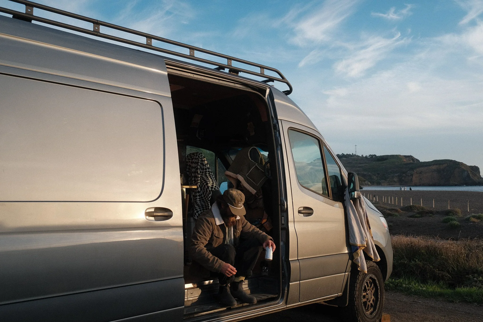 A silver van parked outdoors with the side door open, revealing a man sitting in front of it drinking from a bottle, with another person partially visible in the background. The scene is set in a scenic area with hills or cliffs and a partly cloudy s