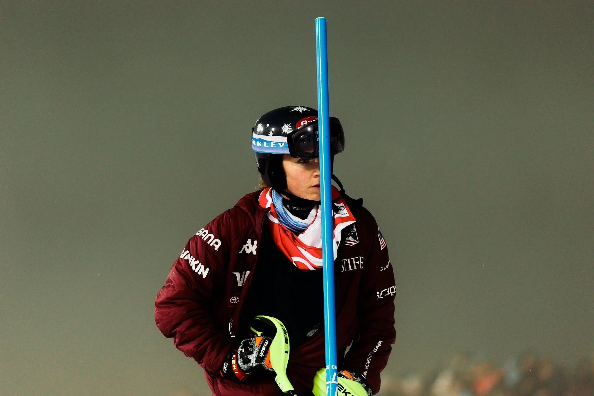 A person dressed in ski gear, holding a blue ski pole and wearing a black helmet with white snowflake designs and goggles, standing against a plain wall.