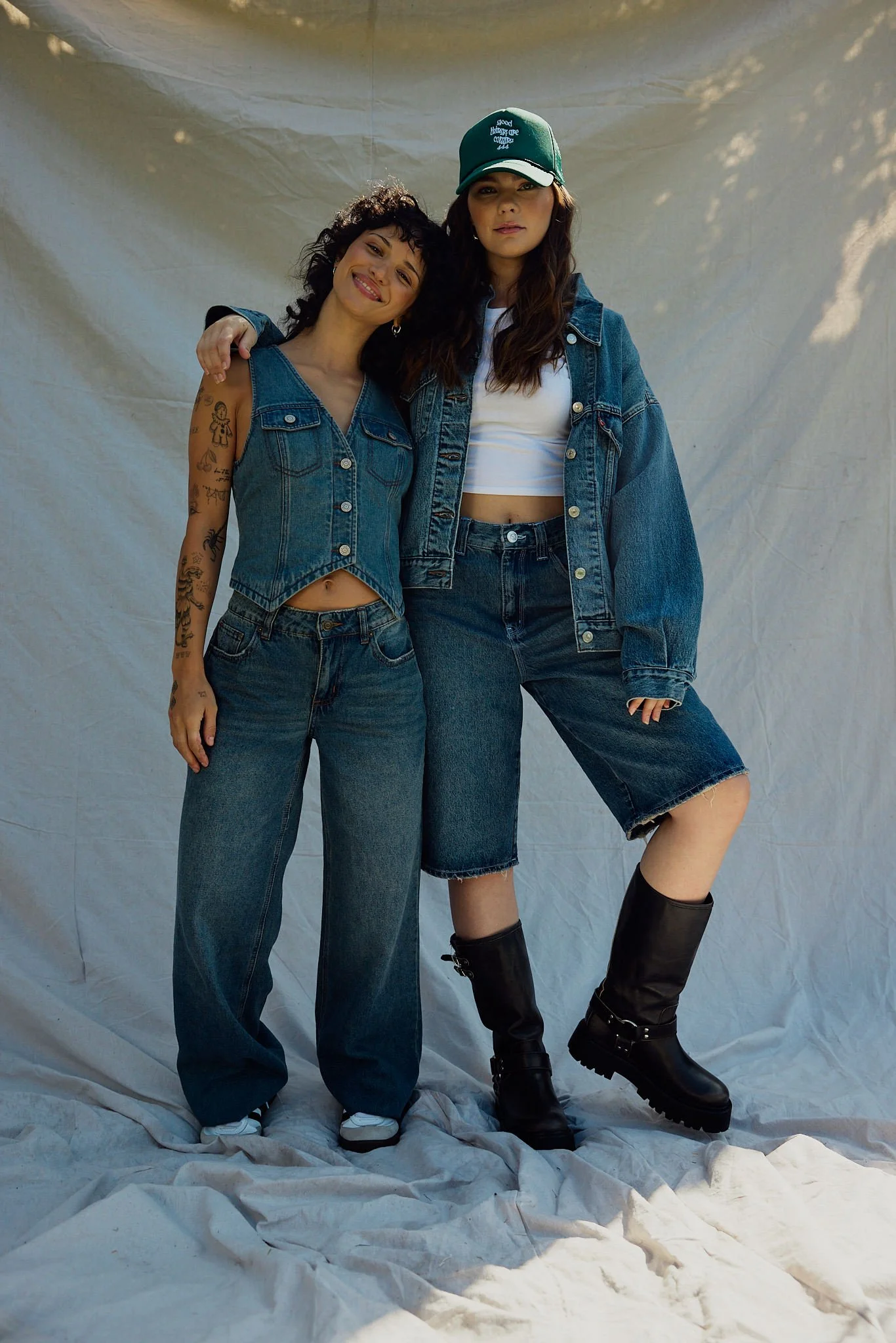 Two young women standing close together, one with curly hair and tattoos on her arm, smiling and wearing a denim vest and jeans, the other with long dark hair, wearing a baseball cap, denim jacket, a white crop top, and jeans, against a plain backdro
